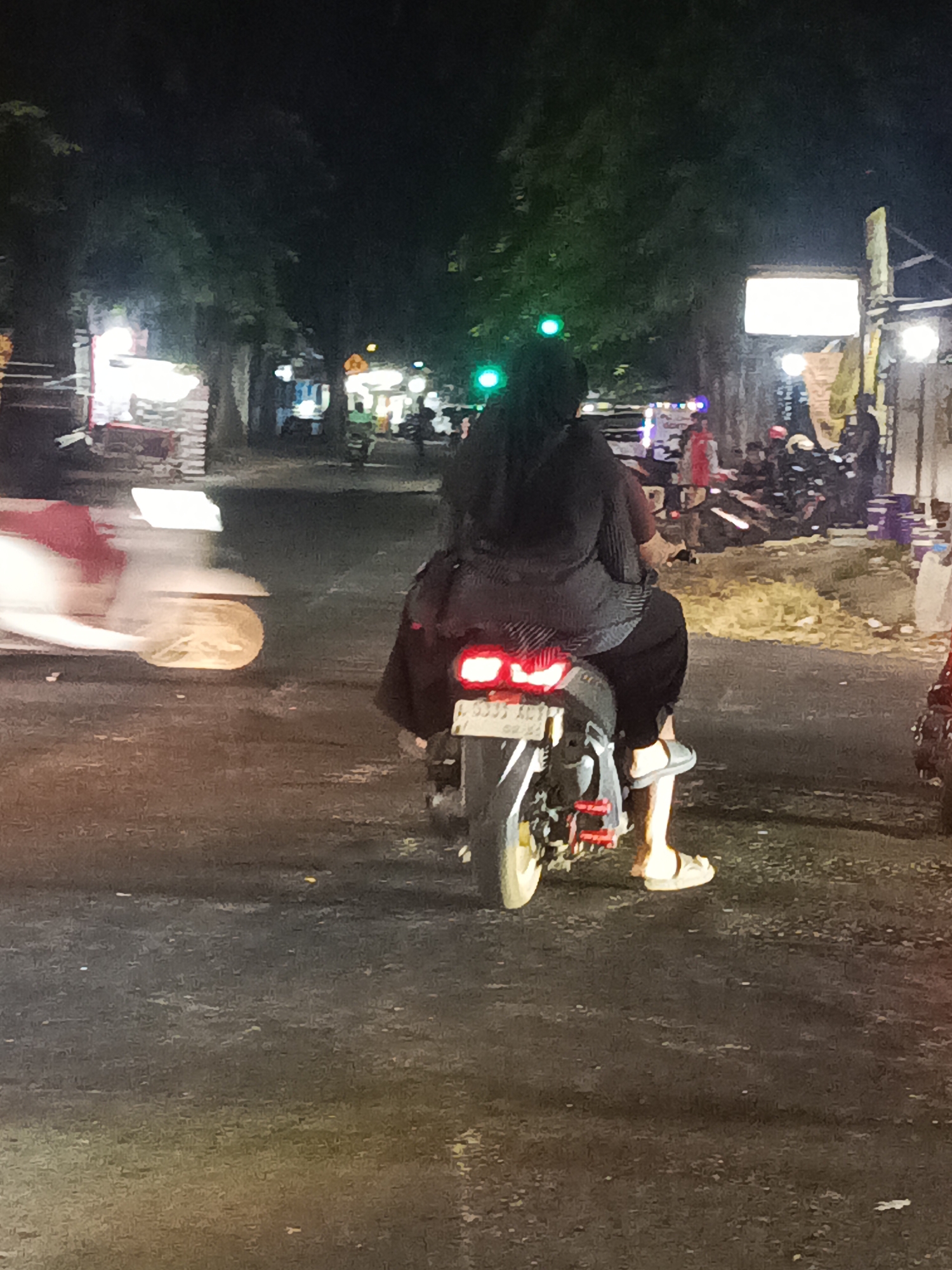 The image captures a nighttime scene on a street in Surabaya, Indonesia, likely near a commercial area given the presence of illuminated stalls or shops in the background. The foreground is dominated by a motorcycle ridden by a person wearing dark clothing and a head covering. The rider's legs are visible, with one leg bent and resting on the footpeg, wearing a light-colored sandal. The motorcycle has a visible license plate, and its taillight is illuminated. To the left of the motorcycle, another vehicle, possibly a scooter, is blurred due to motion, suggesting movement in the scene. The road surface is dark and appears to be asphalt, with some reflections from streetlights. The background shows trees and various lights, indicating it is an urban environment after dark. There are other people and vehicles visible in the background, also illuminated by artificial lights, creating a bustling atmosphere. The overall impression is that of a typical street scene at night in a lively part of the city.