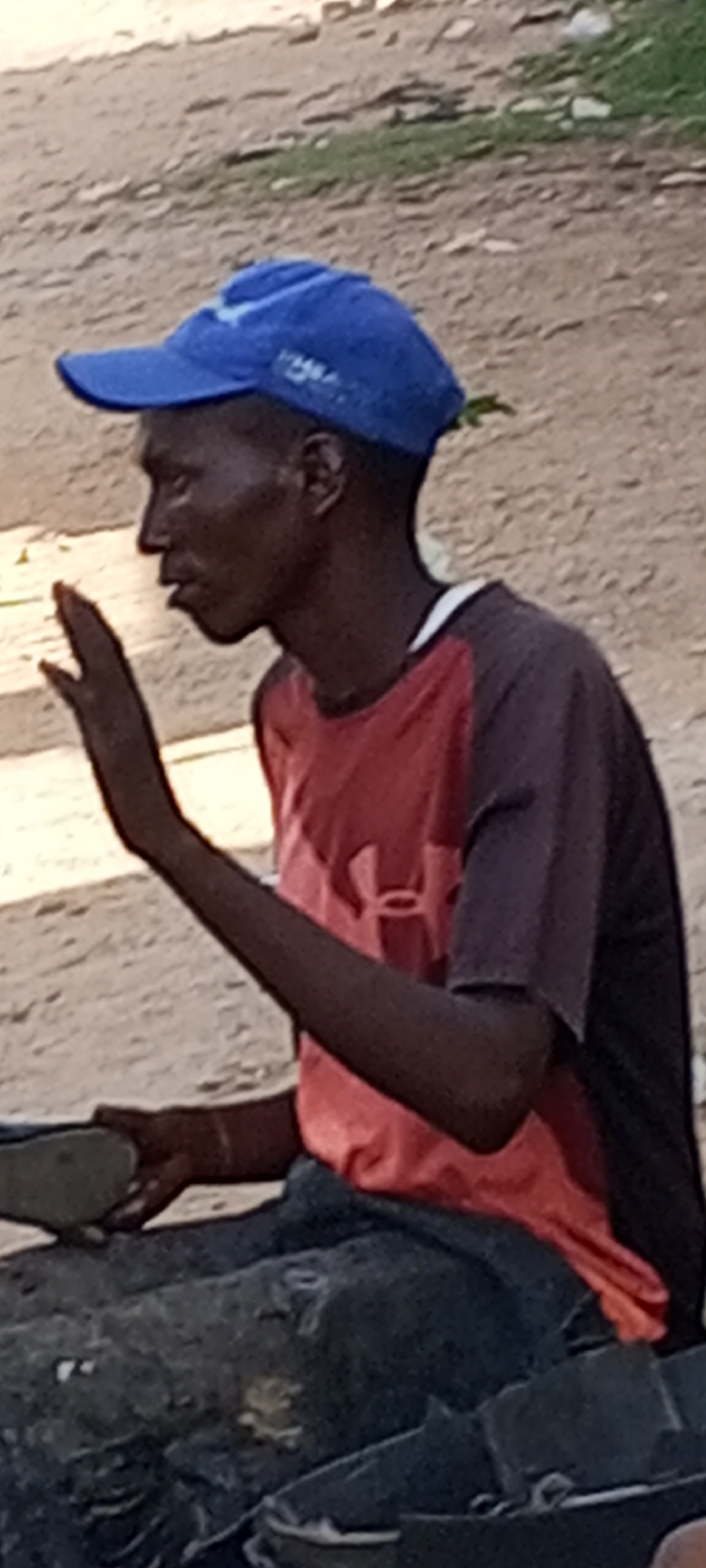 In Sabon Tasha, Nigeria, a dark-skinned man wearing a blue baseball cap and a two-tone short-sleeved shirt (red torso with dark sleeves) sits outdoors. He has a thin build and is gesturing with his left hand raised. In his right hand, he holds a flat object. He sits amidst dark-colored materials, possibly clothing or other soft goods. The background shows a dirt or sandy ground and hints of foliage.