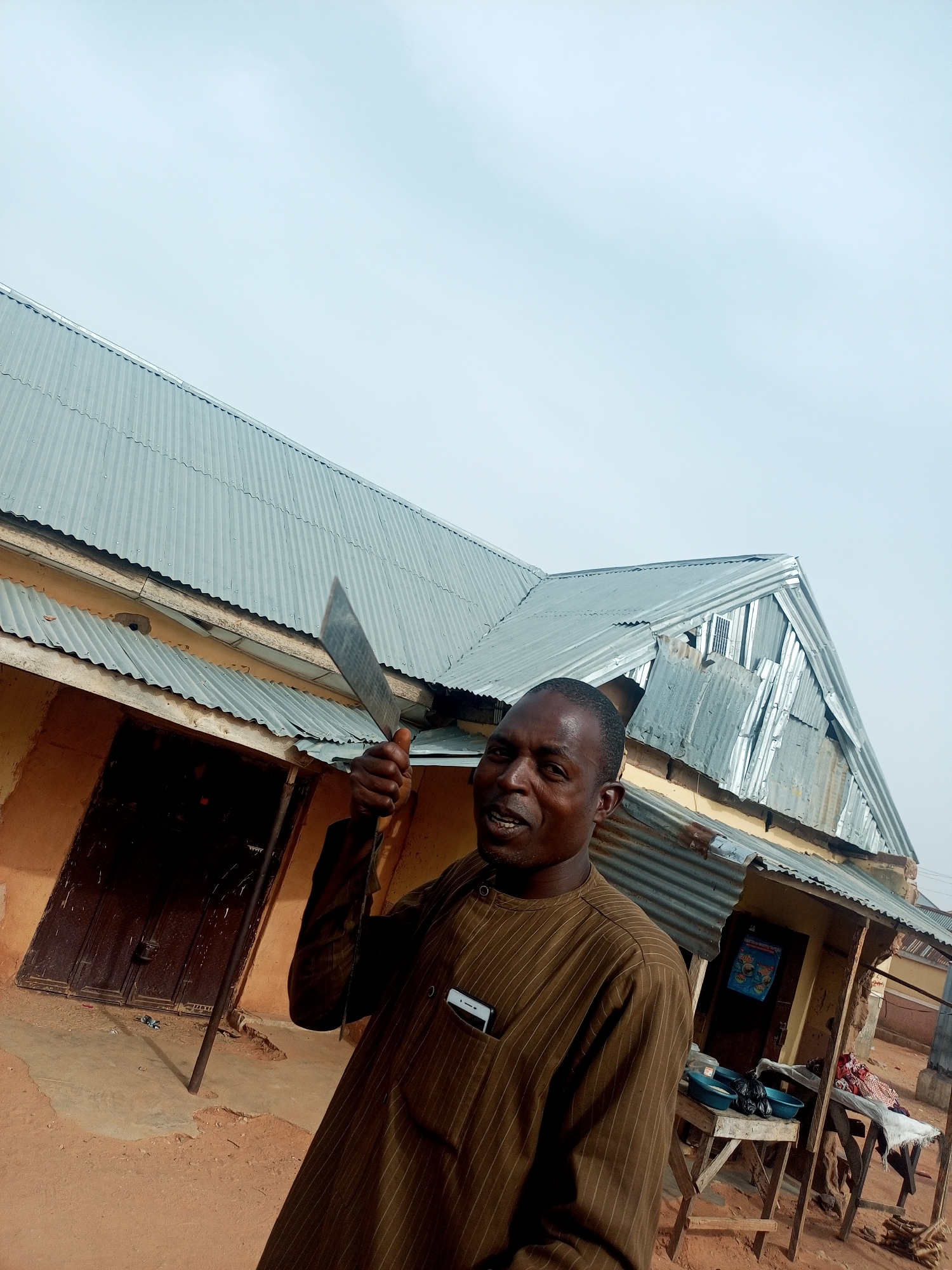 A man stands outdoors, holding a large, silver machete in his right hand, raised near his head. He is wearing a brown, striped, long-sleeved shirt and has a dark complexion. A building with a corrugated metal roof is in the background, partially obscuring the sky. A dark-colored door is visible on the left side of the frame, and a small, weathered structure with a table and containers is on the right. The ground is reddish-brown. The location is Tempure, Nigeria. A white smartphone is visible in the man's pocket.