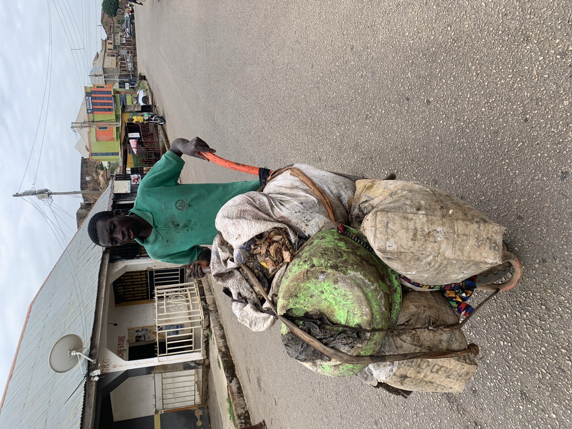 A man in a green shirt is pulling a cart laden with bags and a large, dirty green basin down a paved road. The setting is the city of Jos, Nigeria. Buildings line the road, with one visible establishment identified as "G&G MEDICINE STORE" with a satellite dish attached to its façade. Other multi-storied buildings, some painted in bright colors, are visible further down the road. The sky is overcast. The bags and basin on the cart appear to be filled with refuse.