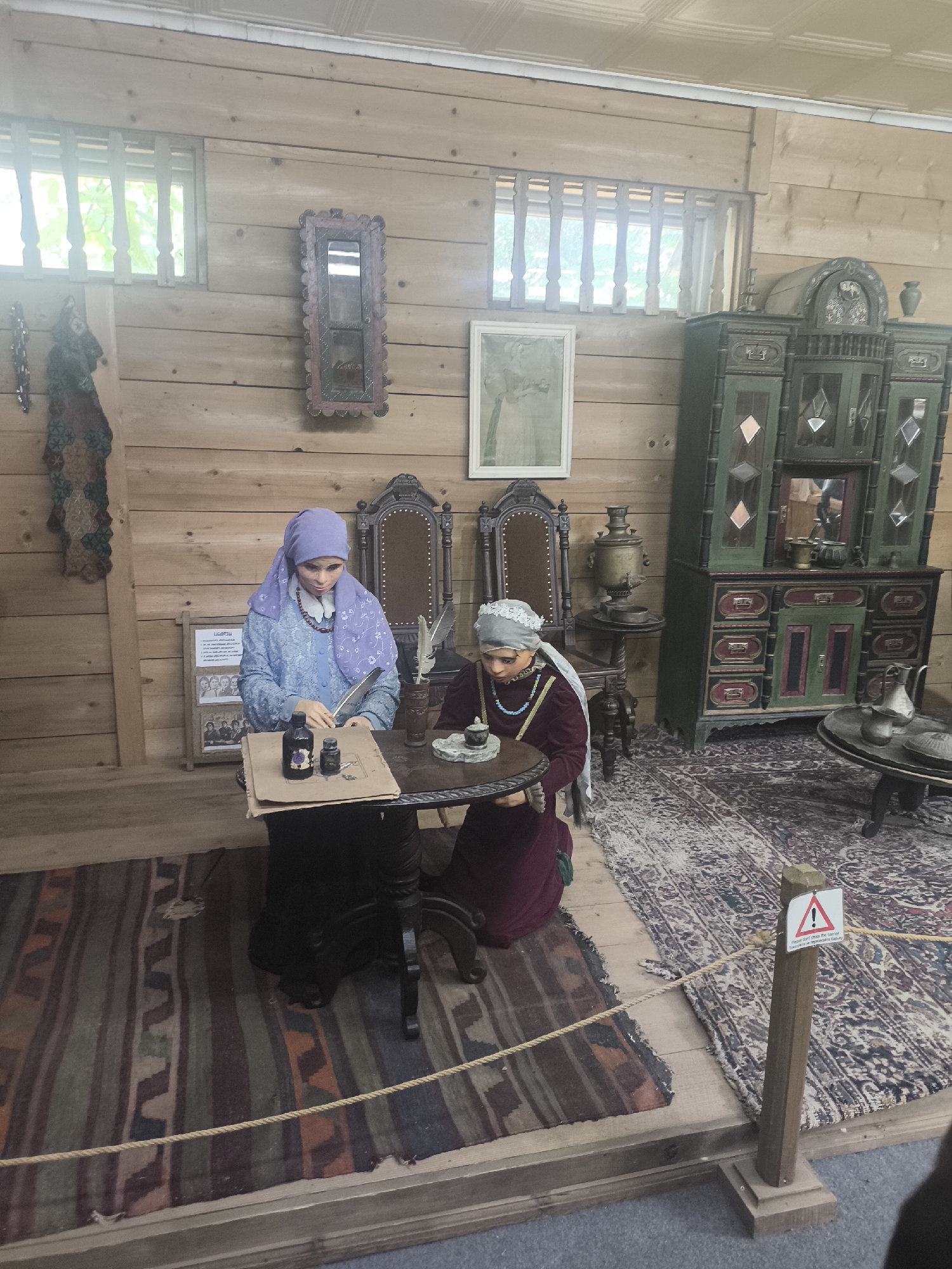 The image shows a diorama of two mannequins dressed in traditional attire, seated at a table in a rustic wooden room. The mannequin on the left, wearing a blue headscarf and dress, is writing with a quill pen, with ink bottles and paper in front of her. The mannequin on the right, in a maroon dress and white head covering, is holding a teacup. Behind the mannequins are various antique items, including chairs, a samovar, and a large, ornate cabinet. A patterned rug covers the floor. The scene is set in the Batumi Ethnographic Museum "Borjgalo", located in Batumi, Georgia, which exhibits traditional Georgian life and culture.