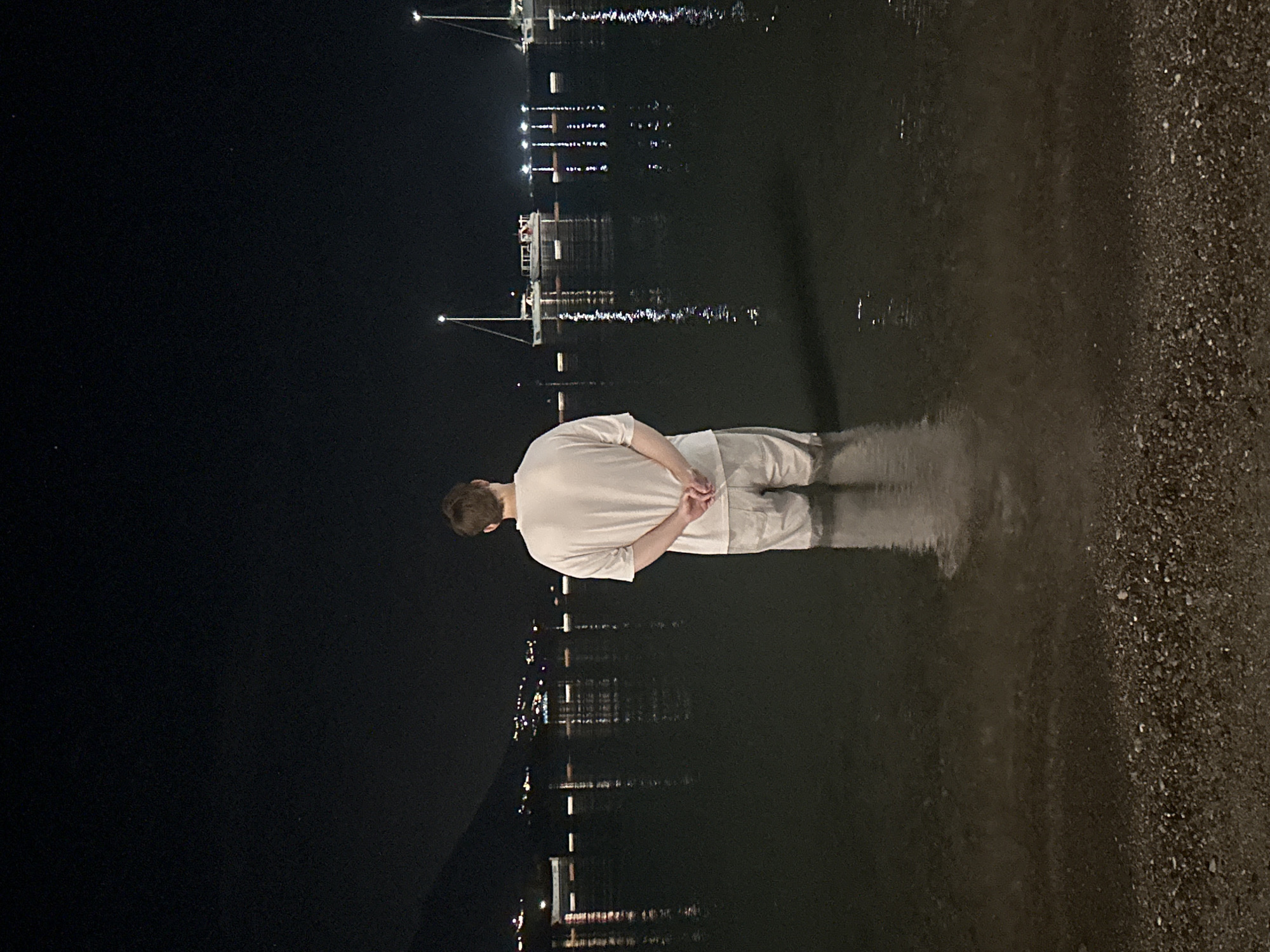 An individual stands waist-deep in dark water at night, facing away from the camera. They are wearing a white t-shirt and light-colored shorts. Their hands are clasped behind their back. The water reflects the lights of a distant city visible on the shoreline, which appears to be Budva, Montenegro. Several boats, including sailboats, are anchored in the water, their lights also reflected. The foreground shows a dark, pebbly beach. The sky is black with a few visible stars.
