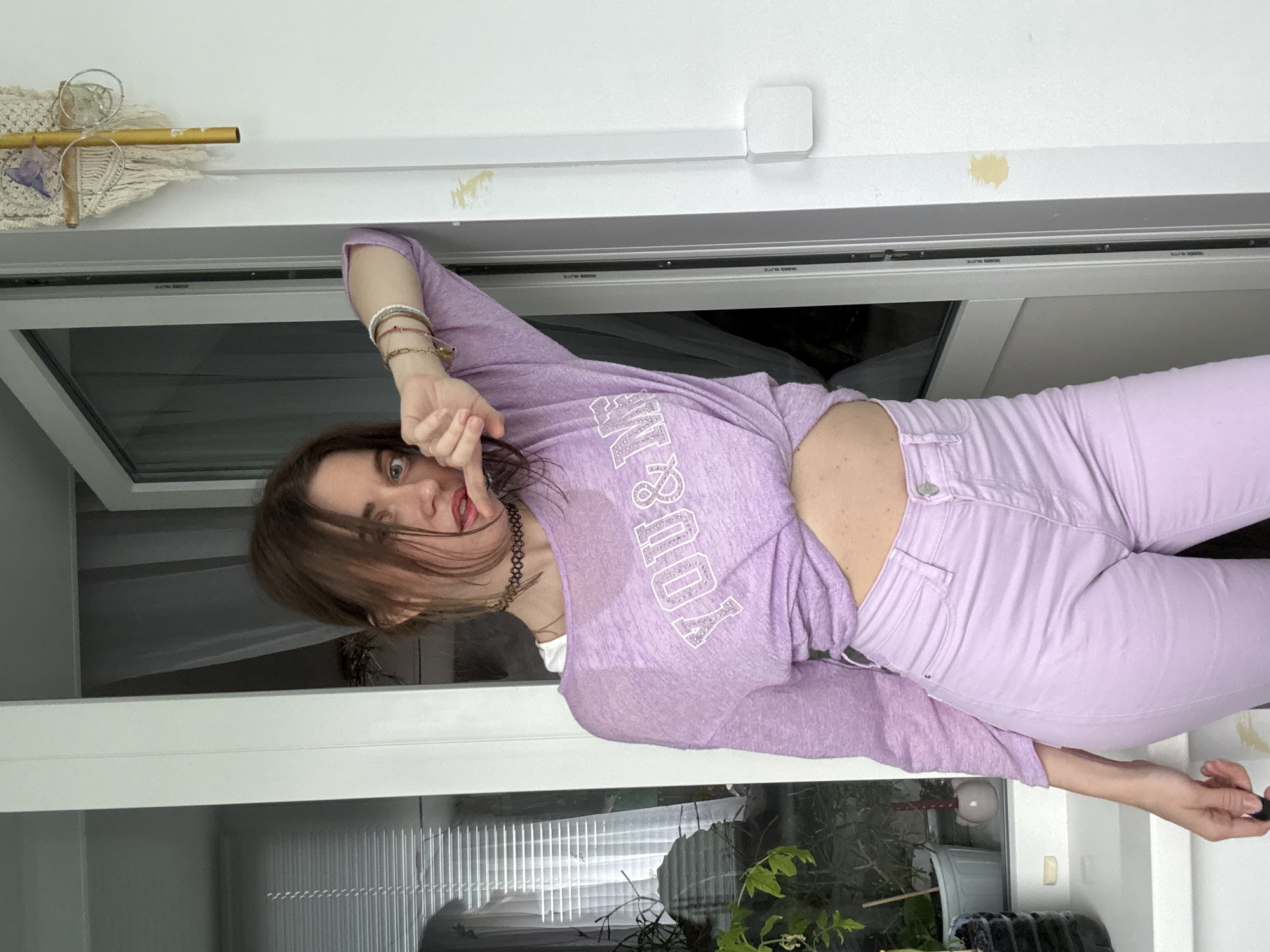 A young woman with brown hair is standing by a doorway, leaning against the frame. She is wearing a purple, cropped long-sleeved shirt with "YOU & ME" in white lettering across the chest, and light purple pants. She has a black choker necklace and several bracelets on her left wrist. Her left arm is bent, with her hand near her mouth, and her right arm is extended, resting on the door frame. The background shows a window with blinds and some plants on a windowsill. The overall setting appears to be indoors, possibly a balcony or a room with a large window.
