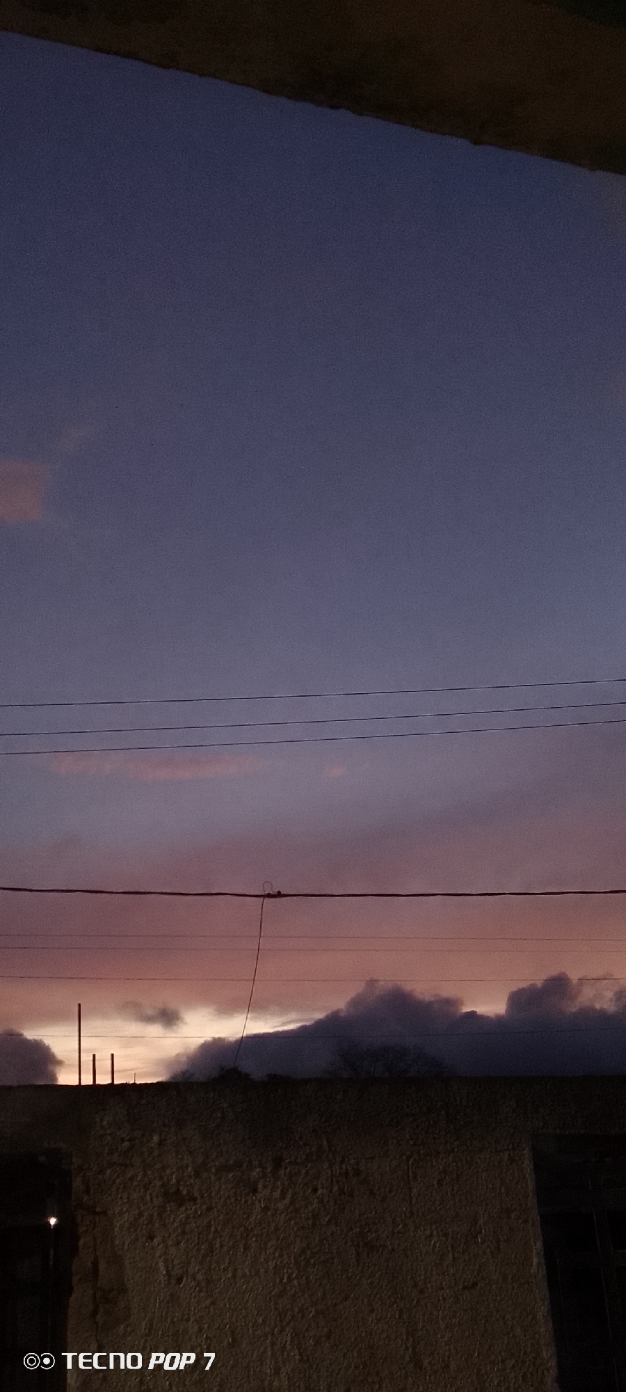 The image captures a dusky scene in Altagracia, Venezuela. An aged, textured wall dominates the lower portion of the frame. Above, several power lines traverse the view, with clouds illuminated by the setting sun partially visible in the space between the power lines and the wall. The sky transitions from a pale orange at the horizon to a deep purple further up. A faint glow suggests the remaining daylight. The "Tecno Pop 7" watermark is visible in the lower-left corner, indicating the phone used to take the image. Some metallic posts are visible on the wall and on top of the roof.