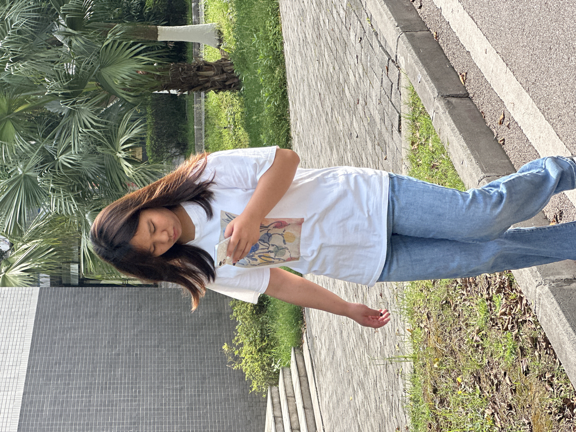 A young woman stands outside on a paved area adjacent to a roadway and a building. She wears blue jeans and a white t-shirt, and holds a phone in her right hand. Her eyes are closed. The pavement appears to be constructed of rectangular pavers, while the roadway is smooth asphalt with a painted white line. There is greenery visible, including small plants, a grassy area, and palm trees. A building with gray tiled walls is also visible. Given the location, the environment could be Jiangjin, China.