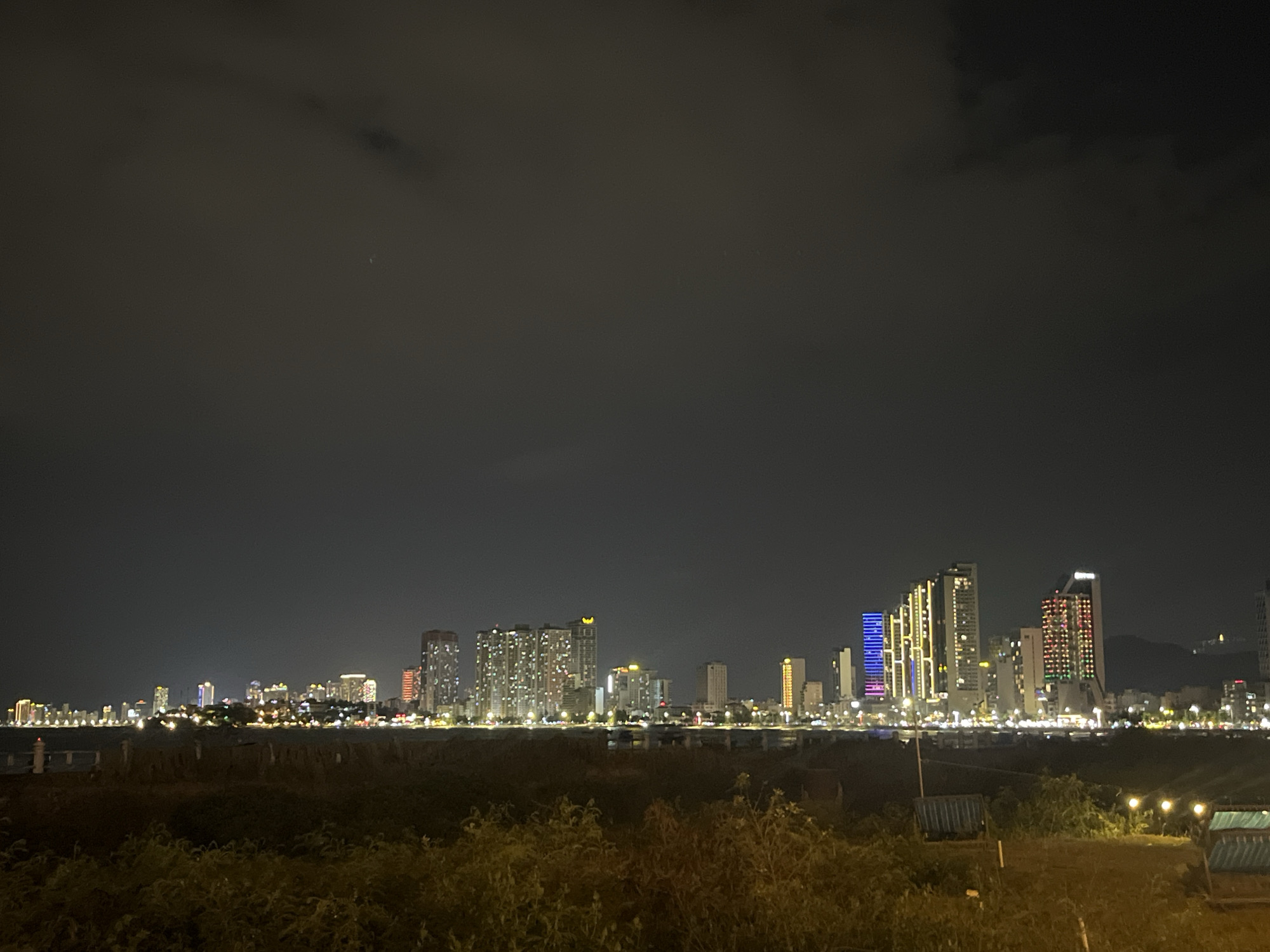 The image depicts a nighttime view of the skyline of Bac Nha Trang Ward, Vietnam. The sky is dark and cloudy. Below the skyline, there's an area of overgrown vegetation, illuminated in the foreground by scattered lights. In the midground, numerous buildings are visible, lit up with an array of colors. The buildings seem densely packed, suggesting a well-developed urban area. The city lights are reflected on what appears to be water, probably the coastline of the sea. Overall, the image captures an urban landscape at night, with the focus on the city's lit-up buildings.