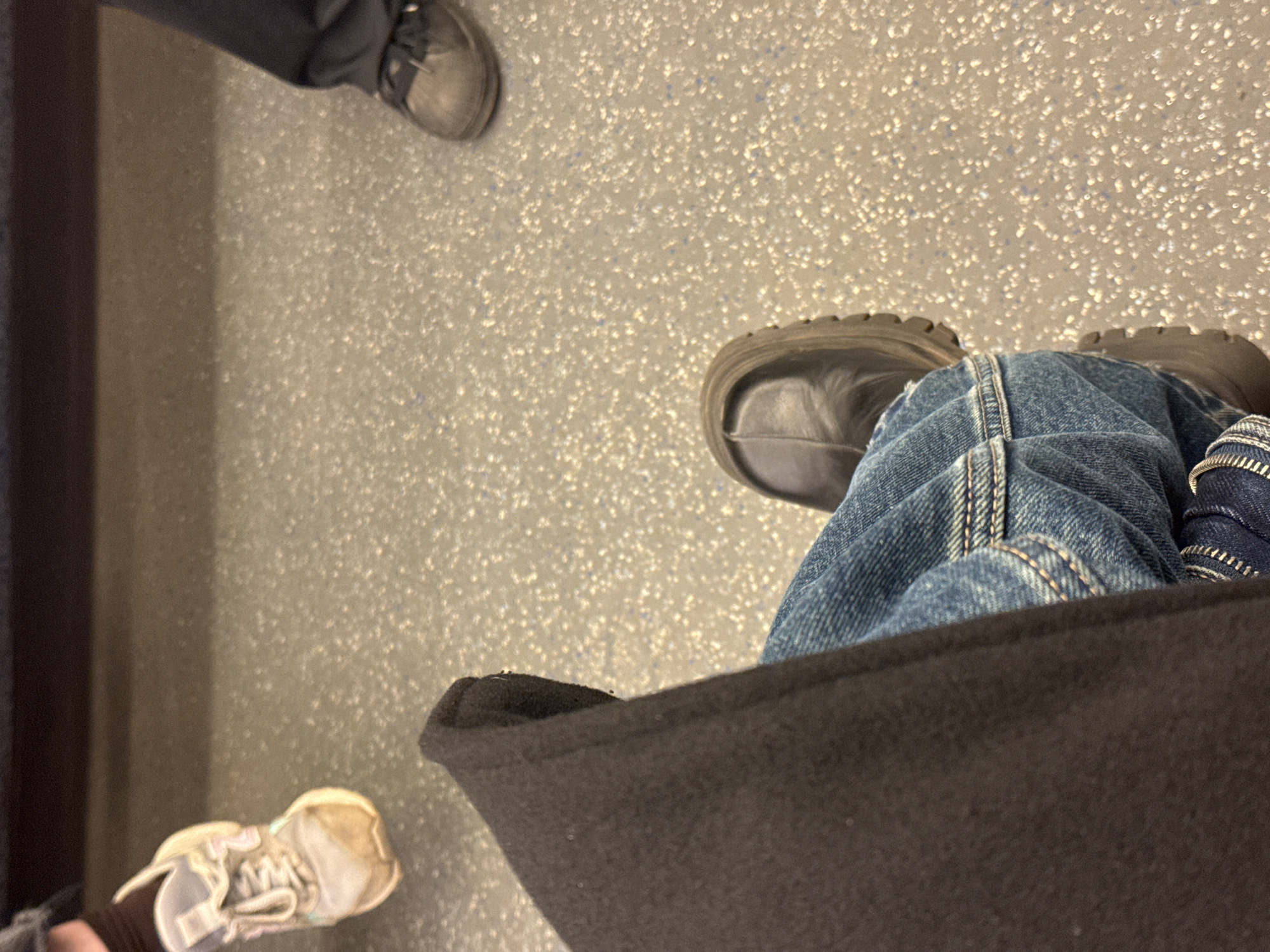 The image shows a ground-level view of a floor with a speckled pattern. Three pairs of feet are visible: one pair wears dark-colored lace-up shoes, a second wears light-colored sneakers, and the third wears dark-colored boots with a lug sole, possibly work boots. The person wearing the boots is also wearing blue jeans and sitting in a black chair. The location is potentially Moscow, Russia, but there is no direct evidence in the image to confirm this, such as recognizable landmarks or Russian text. No specific actions or interactions are evident.