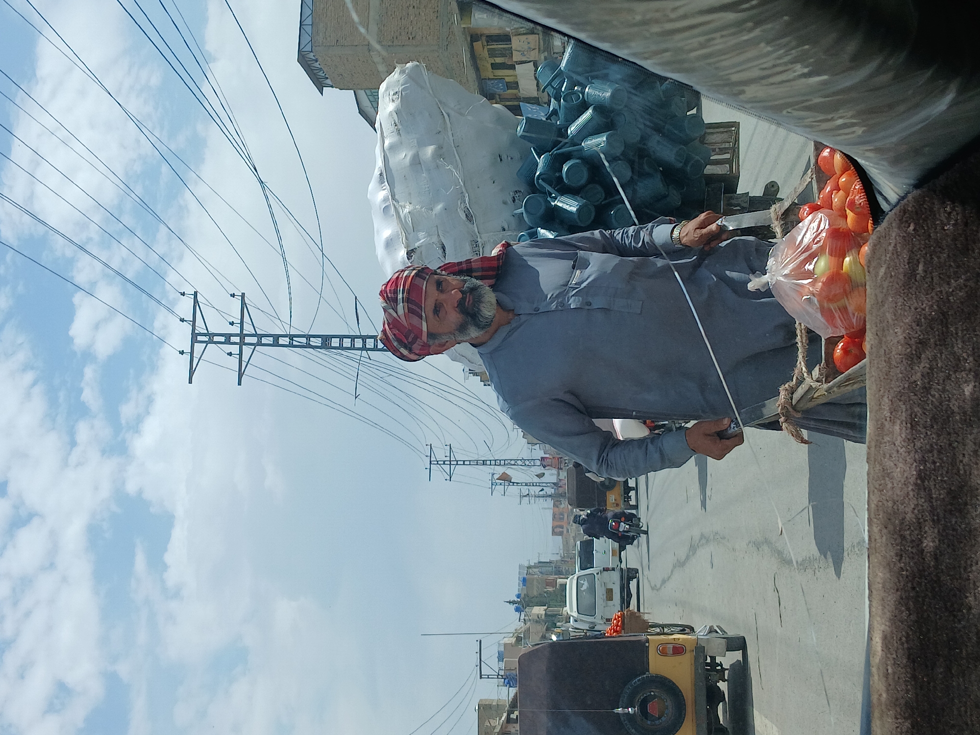 This media file captures a vibrant street scene in Quetta City Tehsil, Pakistan, under a bright, partly cloudy sky. The image, seemingly taken from inside a vehicle, features a man as the central subject, positioned on the right side of the frame.
The man, with a distinguished beard and a traditional red and white checkered turban (likely a pagri or patka style), wears a grey long-sleeved shirt, possibly part of a shalwar kameez. He appears to be engaged in an activity related to the transportation or sale of goods. His left hand is visible, and he wears a metallic watch on his wrist.
Next to the man, under a dark fabric covering, are numerous clear plastic bags overflowing with fresh red and green tomatoes. Behind him, a large, irregularly shaped white bundle, perhaps wrapped goods or material, is visible, along with a stack of dark cylindrical objects that resemble plastic buckets or containers. These items suggest a market setting or a vendor transporting produce and supplies.
The background reveals a bustling street environment. A yellow auto-rickshaw, identified by its spare tire mounted on the back, is visible below the man. Other vehicles, including motorcycles with riders (one wearing a helmet) and white vans, are seen on the road. The street itself is an asphalt surface. Above the street and buildings, numerous overhead power lines crisscross the sky, connected to utility poles, a common sight in urban areas of the region. Buildings with brick and lighter colored facades line the street.
The time of day is clearly daytime, indicated by the bright, natural light. The sky is a mix of blue with scattered white clouds. No visible text is discernable in the image. The scene conveys the daily life and commerce typical of a Pakistani city street.