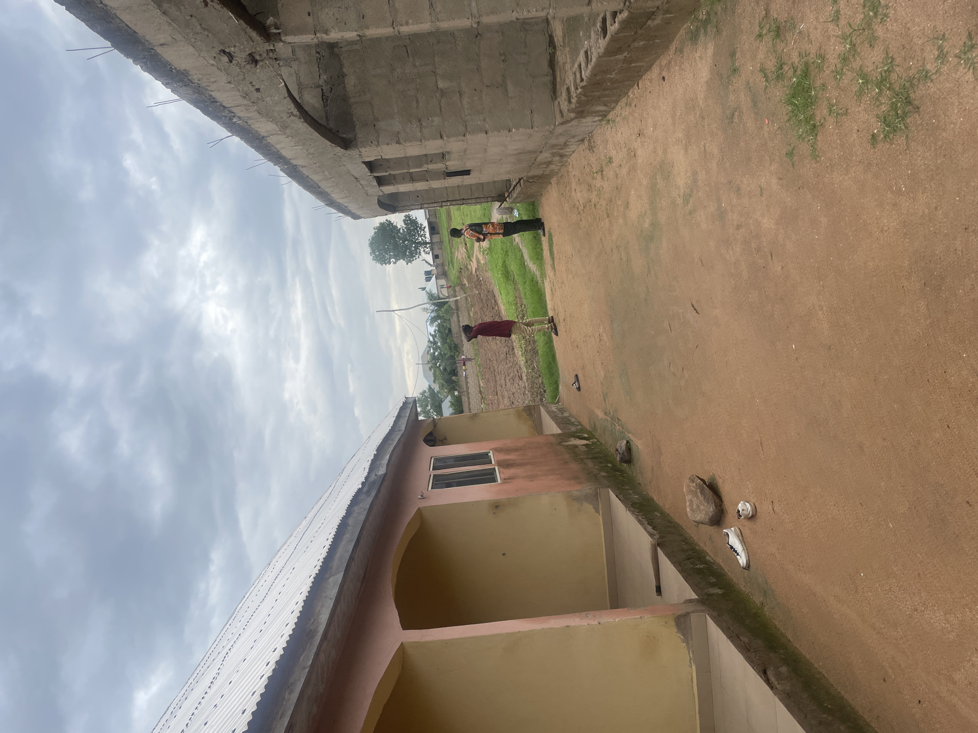 In Jalingo, Nigeria, an outdoor scene captures a dirt pathway between two buildings. To the left, a single-story building with a corrugated metal roof and a pale pink and yellow exterior features arched openings and windows. To the right, a partially constructed building made of concrete blocks stands with exposed rebar at its upper edge and arched window openings. Two men are present on the pathway. One, wearing a red shirt and beige pants, stands facing away from the camera. Another man, in a patterned shirt and dark pants, faces the partially constructed building. In the foreground, a pair of white sneakers rests on the dirt. The sky above is overcast with gray clouds. In the background, agricultural fields and some trees are visible, suggesting a rural or semi-rural setting within the city.