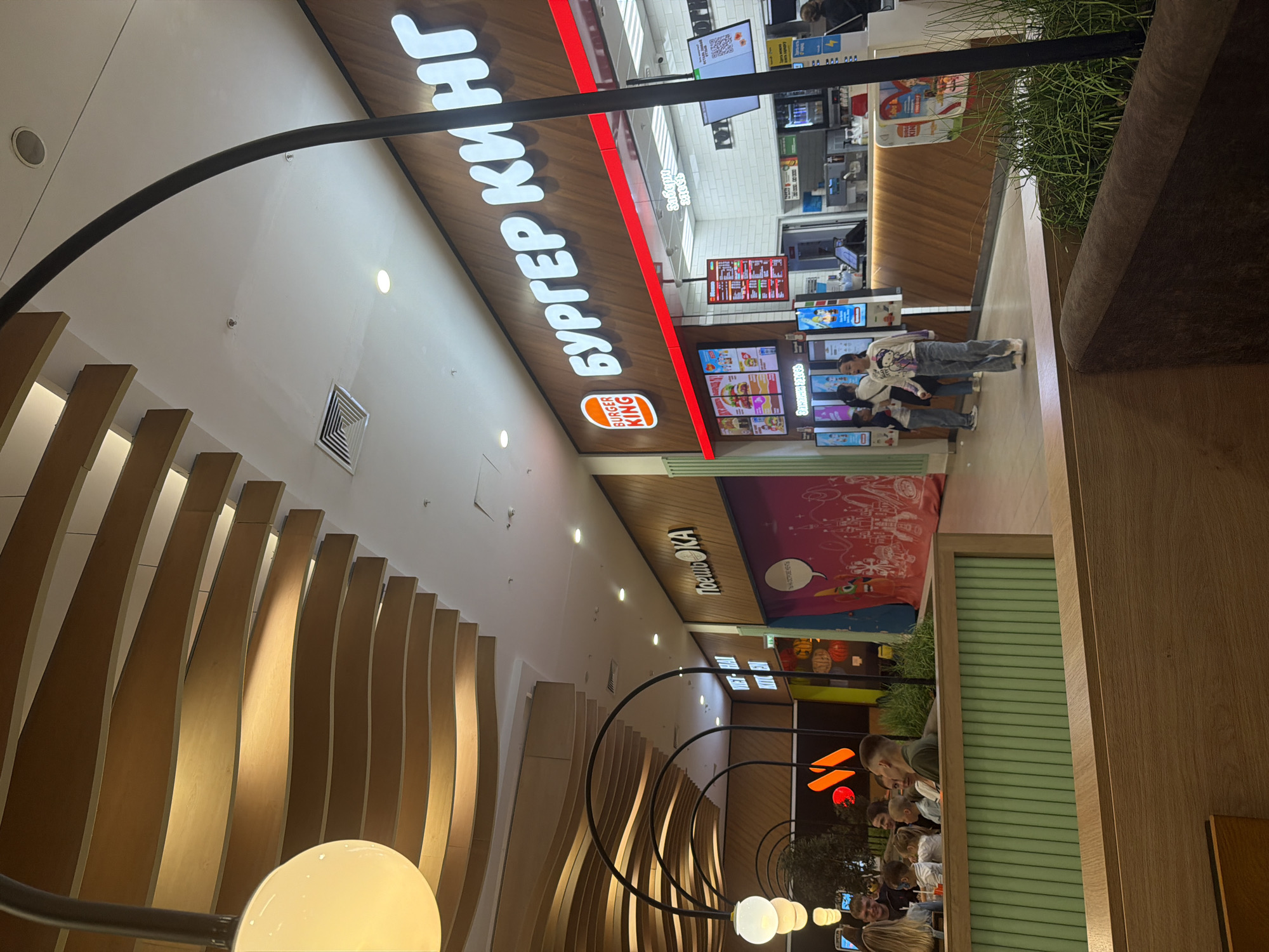 The image depicts a food court in a shopping mall. In the foreground, there are wooden tables and green partitioned seating areas. A group of people are seated at one of the tables, seemingly enjoying a meal. In the background, several food establishments are visible, including a Burger King with its distinctive logo and signage. There are also digital menus displaying food items. People are seen ordering and waiting for their food. The ceiling features a modern, curved wooden design with ambient lighting. The overall environment suggests a bustling and contemporary dining space.