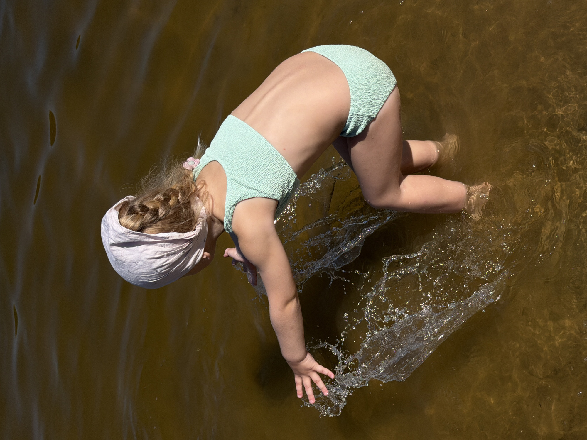 A young child wearing a light green textured swimsuit and a pink headscarf is in shallow, murky brown water. The child is on their hands and knees, with their head and torso bent downwards. Their right arm is extended, creating a splash of water. Their legs are bent, with their feet submerged in the water. The child's blonde hair is braided and visible under the headscarf, with a small pink flower accessory. The scene appears to be a natural body of water, possibly a lake or river, given the murkiness and ripples on the surface. The lighting suggests it is daytime, likely sunny, as indicated by the bright highlights on the water splashes. No text is visible in the image.