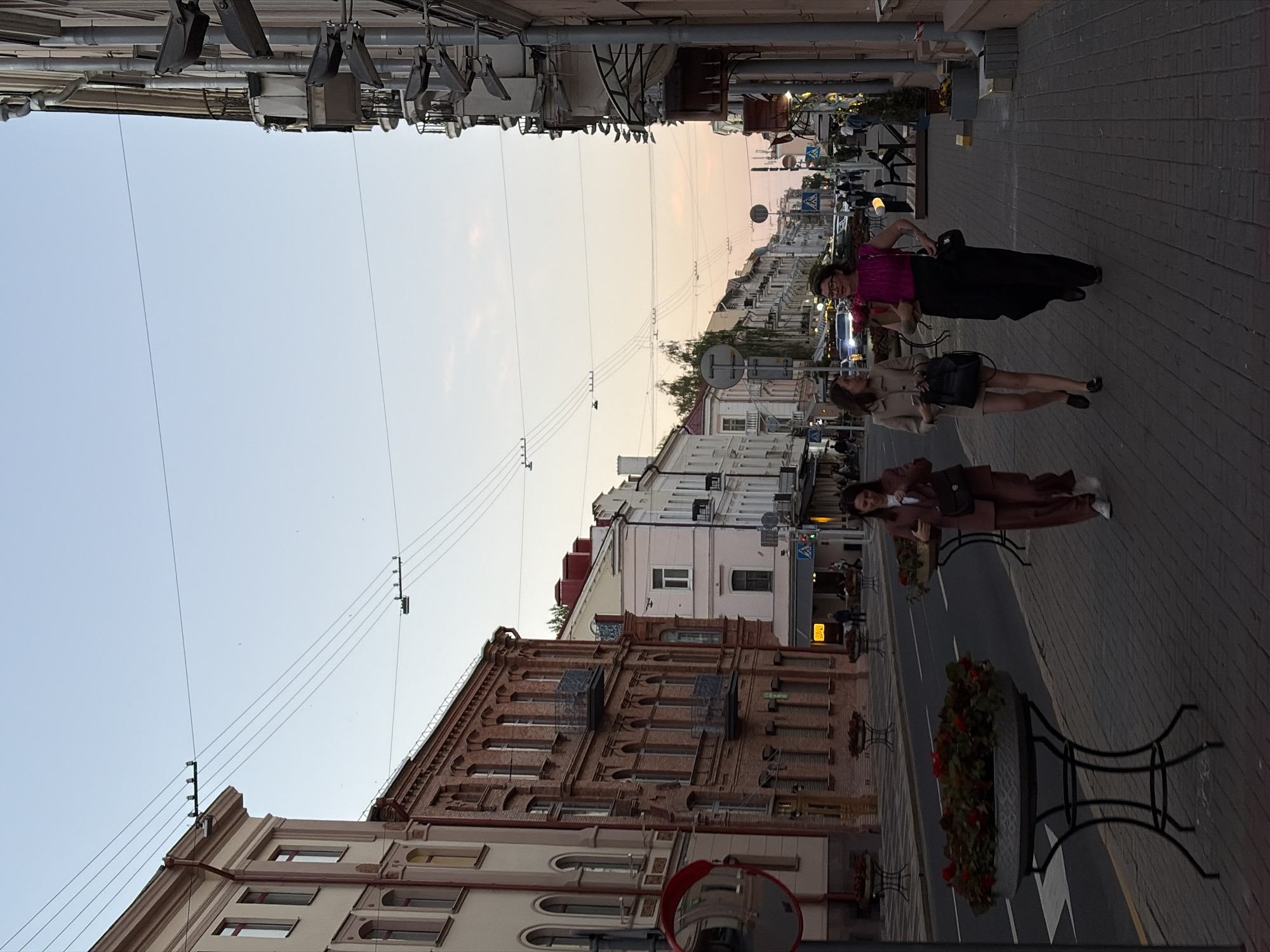 The image captures a bustling street scene in Minsk, Belarus, during dusk or early evening. The sky, visible at the top, transitions from a clear light blue to a soft orange and pink haze near the horizon, indicative of a fading sunset.
The street is lined with various multi-story buildings, showcasing a mix of architectural styles. On the left side, a prominent building features intricate red brickwork, arched windows, and decorative cornices, contrasting with a lighter-colored facade closer to the foreground. Overhead, numerous power lines or tram lines stretch across the frame, crisscrossing the sky.
The right side of the image features a wide paved sidewalk and a roadway with white lane markings. Several dark planters overflowing with vibrant red flowers are spaced along the pavement. Three women are present on the sidewalk:
1. In the foreground, a woman with glasses wears a bright pink top and a long black skirt, standing with her hands on her hips, observing the surroundings.
2. Slightly behind her, another woman is dressed in a light beige jacket and a darker skirt or shorts, carrying a black handbag.
3. Further back, a third woman wears a stylish brown suit with white sneakers, standing near one of the flower planters.
In the middle and far background, more buildings line the street, some with illuminated windows or signs. Visible details include streetlights, a blue and white traffic sign, and faint outlines of distant vehicles. A round convex mirror is partially visible in the bottom-left foreground, reflecting a portion of the nearby architecture. The overall atmosphere is relaxed, depicting a typical urban evening setting. No discernible text is clearly readable on any signs.