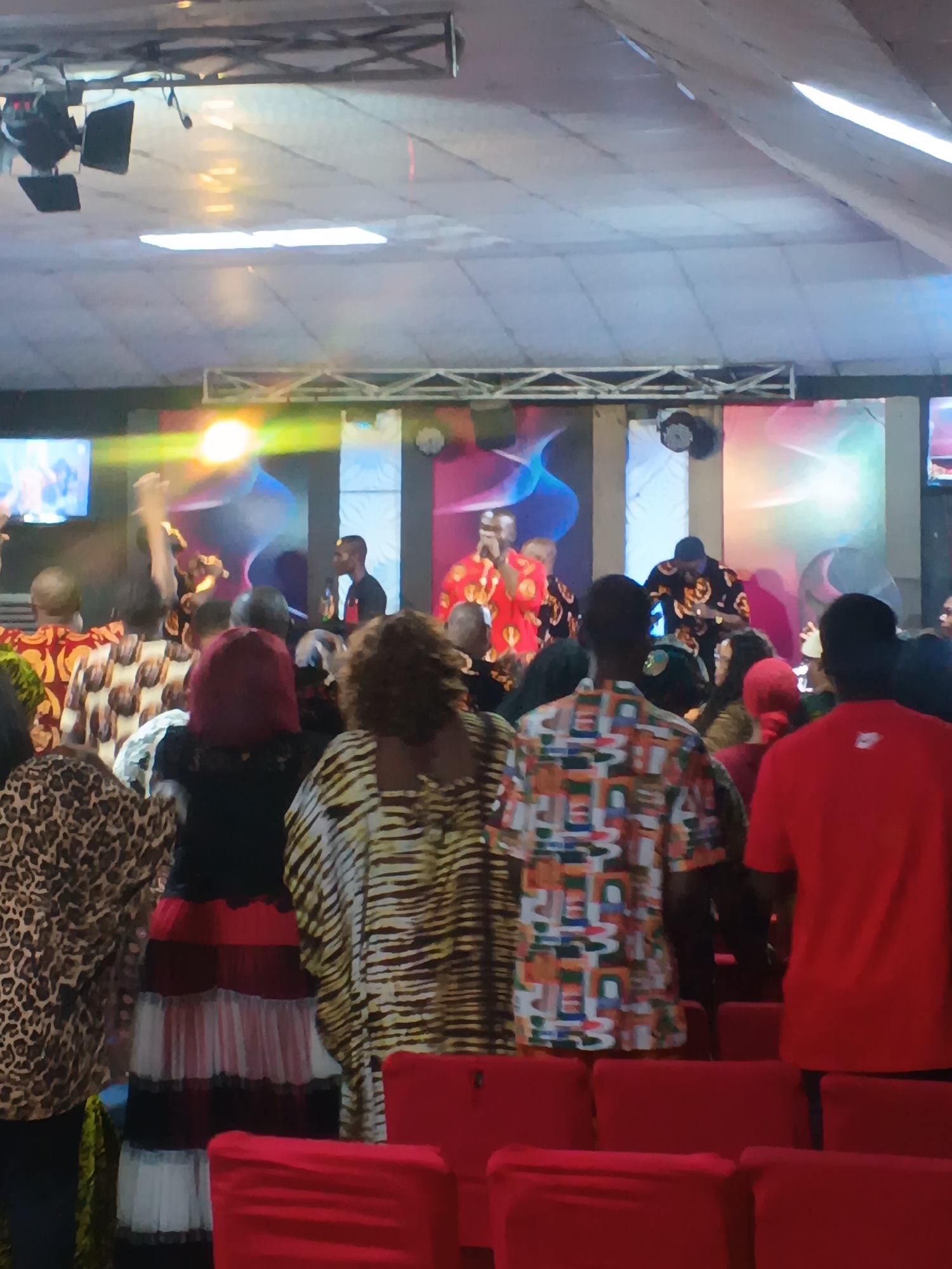 The image captures an indoor scene, likely a church or event hall, with a group of people gathered. The foreground is dominated by rows of red chairs, some covered with fabric. A few individuals are visible from the back, wearing brightly colored and patterned clothing, including a leopard print dress and a dress with horizontal black, red, and white stripes.
In the middle ground and background, a stage area is set up with a backdrop featuring abstract, colorful graphics. Several individuals are on the stage, appearing to be performers or speakers. One person in the center is wearing a vibrant, patterned shirt and appears to be engaged in an activity. Another person on the left is wearing a red outfit with intricate patterns. A man on the right is speaking into a microphone. Lighting equipment is visible above the stage. The overall atmosphere suggests a lively gathering, possibly a religious service or a musical performance. The presence of the church name "Grace Foundation International" on a visible banner, combined with the context of a gathering and stage setup, strongly indicates the location is Grace Foundation International Church in Nkpor, Nigeria.