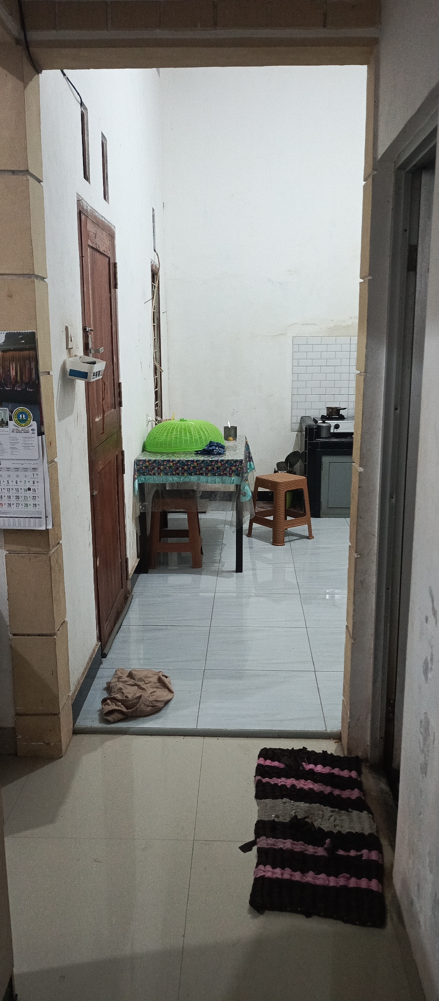 The image shows a narrow hallway leading into a room that appears to be a kitchen area. On the left, a wooden door is visible, with a calendar hanging on the wall next to it. The hallway floor is tiled. The room beyond contains a table with a green cover and a small candle, with two stools on either side. A fabric item is lying on the floor in front of the table. In the background, a small kitchen counter with a stove and white tiled backsplash is visible. A striped rug lies on the floor of the hallway, near the entrance to the room. The scene suggests a residential interior in Arjasa, Indonesia.