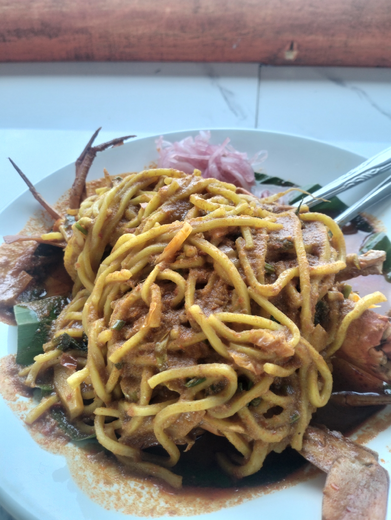 A close-up shot shows a plate of noodles with crab, served in Lancok, Indonesia. The noodles are yellow and curly, coated in a dark sauce. Several pieces of crab, including legs and claws, are visible beneath and around the noodles. A garnish of thinly sliced red onions and green leaves is present on the side. The plate rests on a white surface, likely a table, with a textured wooden background in the upper portion of the frame. The lighting is natural, suggesting an indoor setting with ambient light.