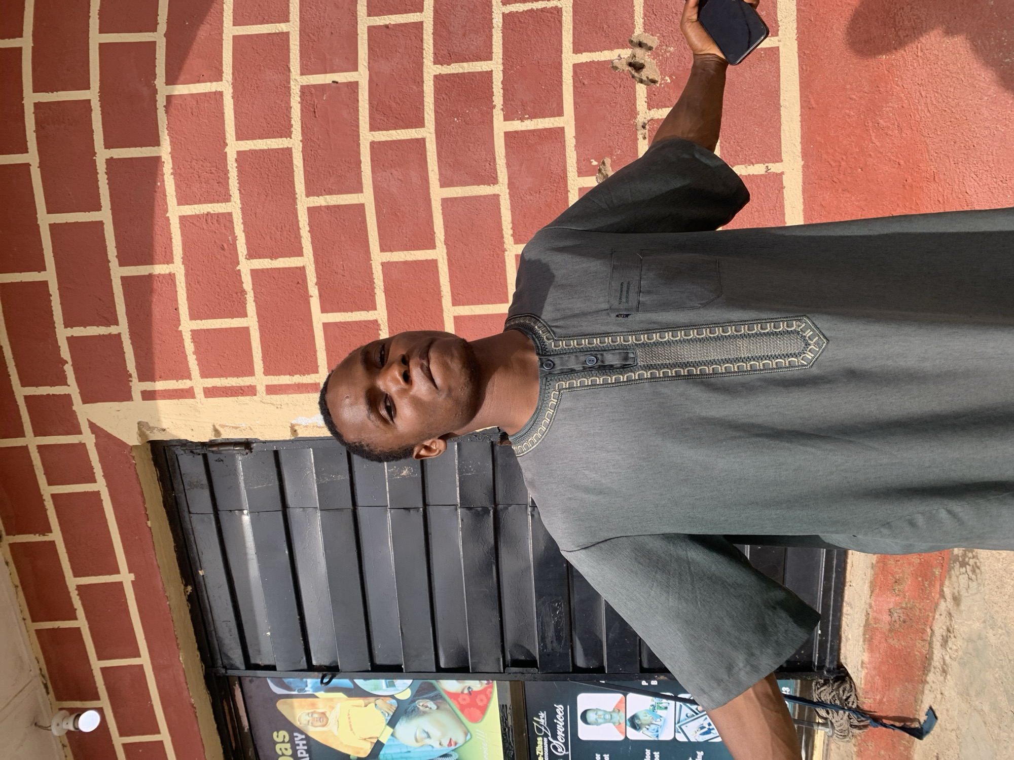 A young man is pictured outdoors, holding a dark-colored smartphone in his right hand. He is wearing a gray, long-sleeved tunic with ornate stitching around the neckline and down the front. The tunic has a button closure. He is standing in front of a red brick wall with yellow mortar. To his left, there is a black metal door or gate with a ribbed surface. Below the door, a sign is partially visible, displaying images of people and text. One piece of visible text reads "raph". The lighting suggests it is daytime, with shadows cast on the brick wall. The man's expression appears neutral. The location is identified as Jos, Nigeria.