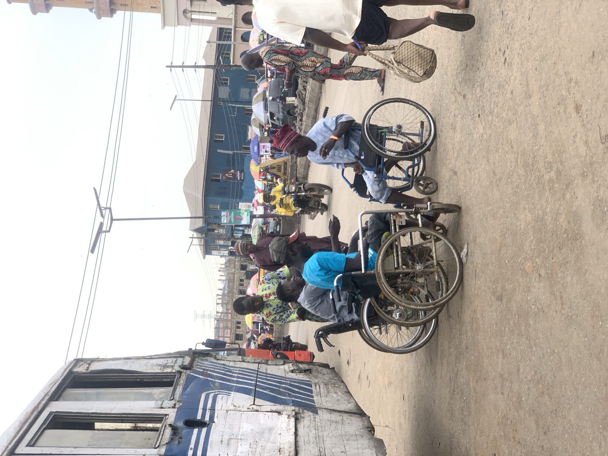 The image shows a street scene in Ikorodu, Nigeria. Several men in wheelchairs are moving along the road; some are wearing traditional Nigerian clothing, including a man with a maroon hat. A man walks past them carrying a wicker basket. Vehicles and buildings are visible in the background. The road is paved. An old, blue and white bus is parked along the side of the road. Power lines and utility poles are overhead.