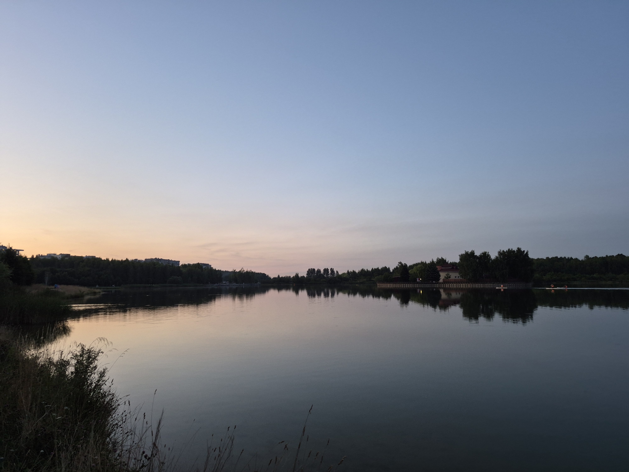 This is a landscape photograph of a tranquil lake at dusk, likely in the city of Minsk, Belarus. The sky transitions from a soft, pale blue at the top to a delicate peachy-pink hue near the horizon, indicating the last moments of daylight. The water's surface is calm, reflecting the colors of the sky and the silhouettes of trees and buildings along the far bank. On the left side of the image, the shore is lined with tall grasses and brush in shadow. The middle ground of the scene features a dense line of dark green trees, broken by some buildings on the left bank, suggesting a suburban or park area. On the right side of the lake, a building with a reddish-brown roof is visible amidst the trees, and two small figures in what appear to be kayaks are present on the water, moving from right to left. The overall atmosphere is peaceful and serene.