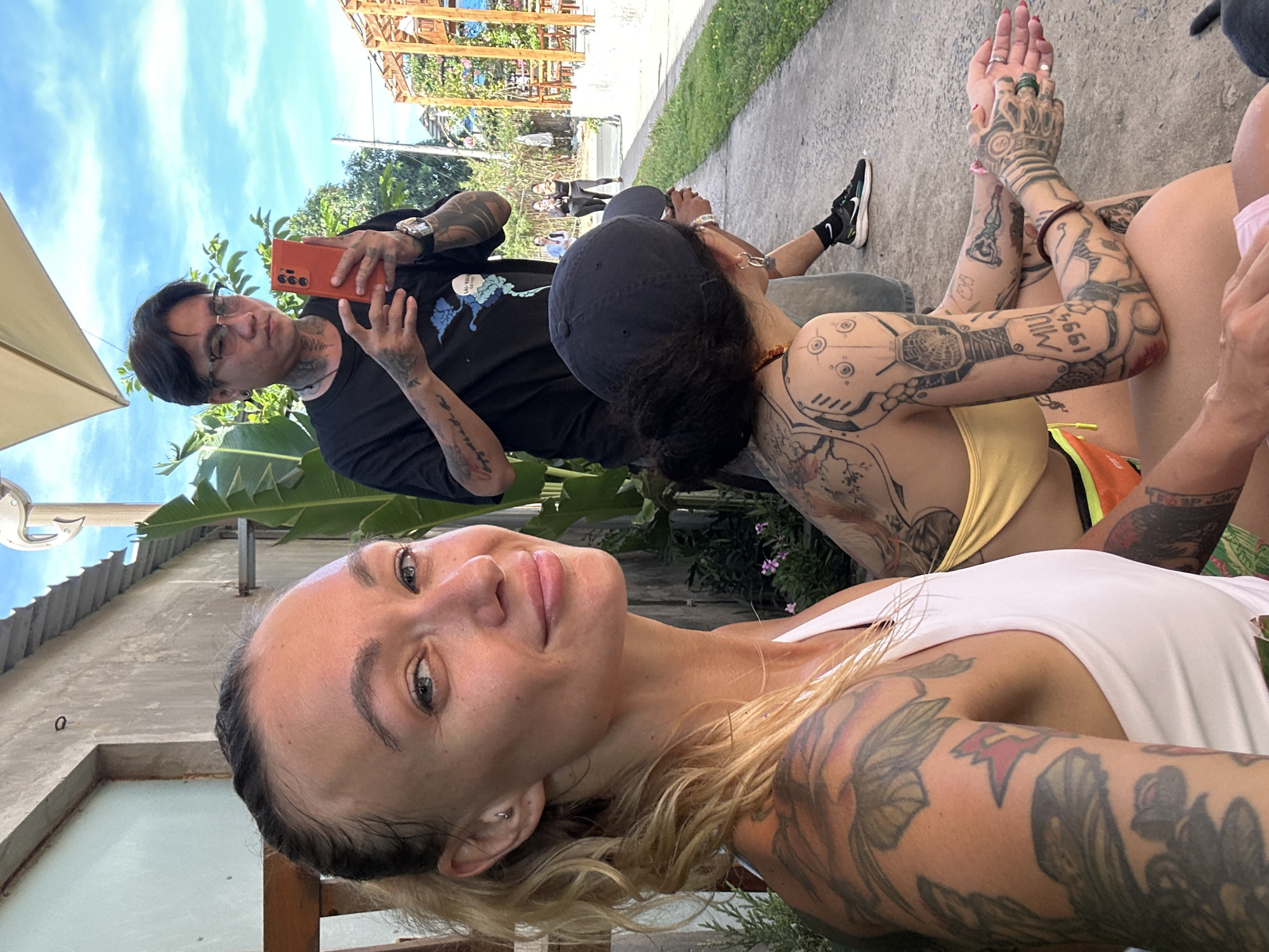 In Phu Thuy Ward, Vietnam, three individuals are depicted in an outdoor setting. In the foreground, a woman with blonde hair tied back is positioned on the left, facing the camera with a slight smile. She is wearing a white tank top and has extensive tattoos covering her arms and upper chest. To her right, partially obscured, is another individual, viewed from the back, wearing a dark baseball cap and a yellow top. This person also has numerous tattoos, particularly on their arms and back. In the background, a man wearing glasses, a black t-shirt, and dark pants is holding an orange smartphone. He appears to be taking a photo or video. The environment is outdoors, with a concrete pathway in the foreground and a building with a corrugated roof visible on the left. Green foliage and trees are present, and the sky is blue with some clouds. In the distance, a wooden structure and other people can be seen. The overall scene suggests a casual gathering or outing in a moderately populated area.