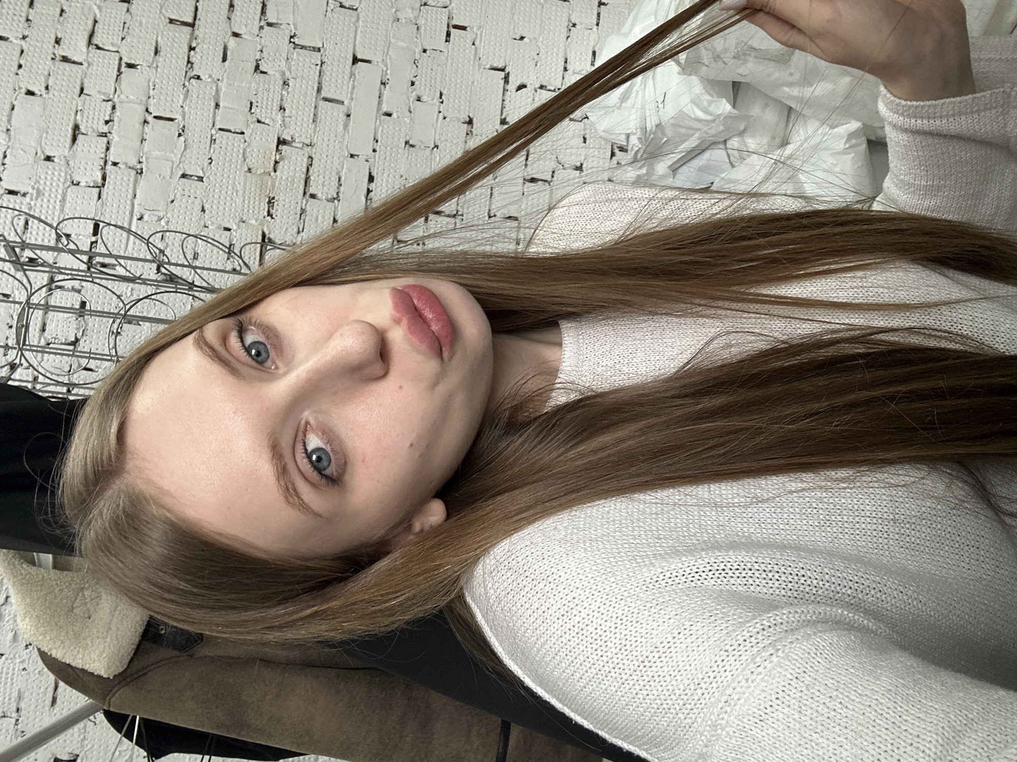 A young woman with light skin, light blue eyes, and long, light brown hair is positioned in the foreground. She is wearing a white knitted sweater and holding a strand of her hair up with her hand. The background consists of a white, textured wall reminiscent of brickwork. To the left, there is a metal clothing rack partially visible with some clothing including a coat or jacket with a cream-colored shearling collar. Other elements in the background are out of focus. The location in the city of Chaikovsky, Russia is not discernible from the provided image.
