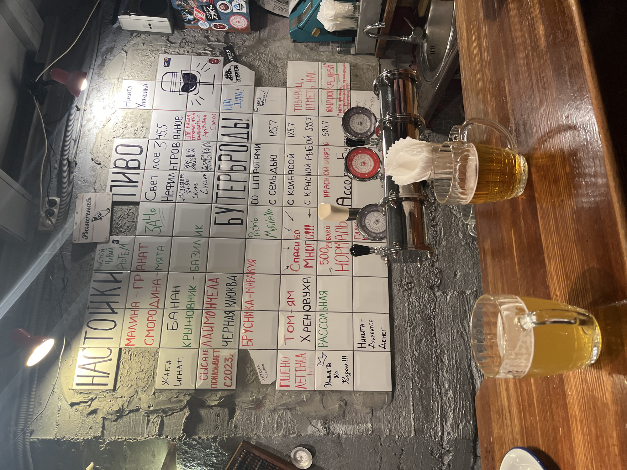 A bar in an unknown city, possibly in Russia, features a wall menu written in Russian on white square tiles. The menu lists various alcoholic drinks, including infused spirits ("NACTOйKИ"), beer ("ПИBO"), and sandwiches ("БУTEPБPOЛЫ"). Prices are indicated next to the menu items. There are two beer mugs filled with amber-colored beer on the wooden bar top, positioned to the right of a beer tap system. The wall and the surrounding area have a textured, possibly concrete or plaster finish. The overall lighting in the space is dim.