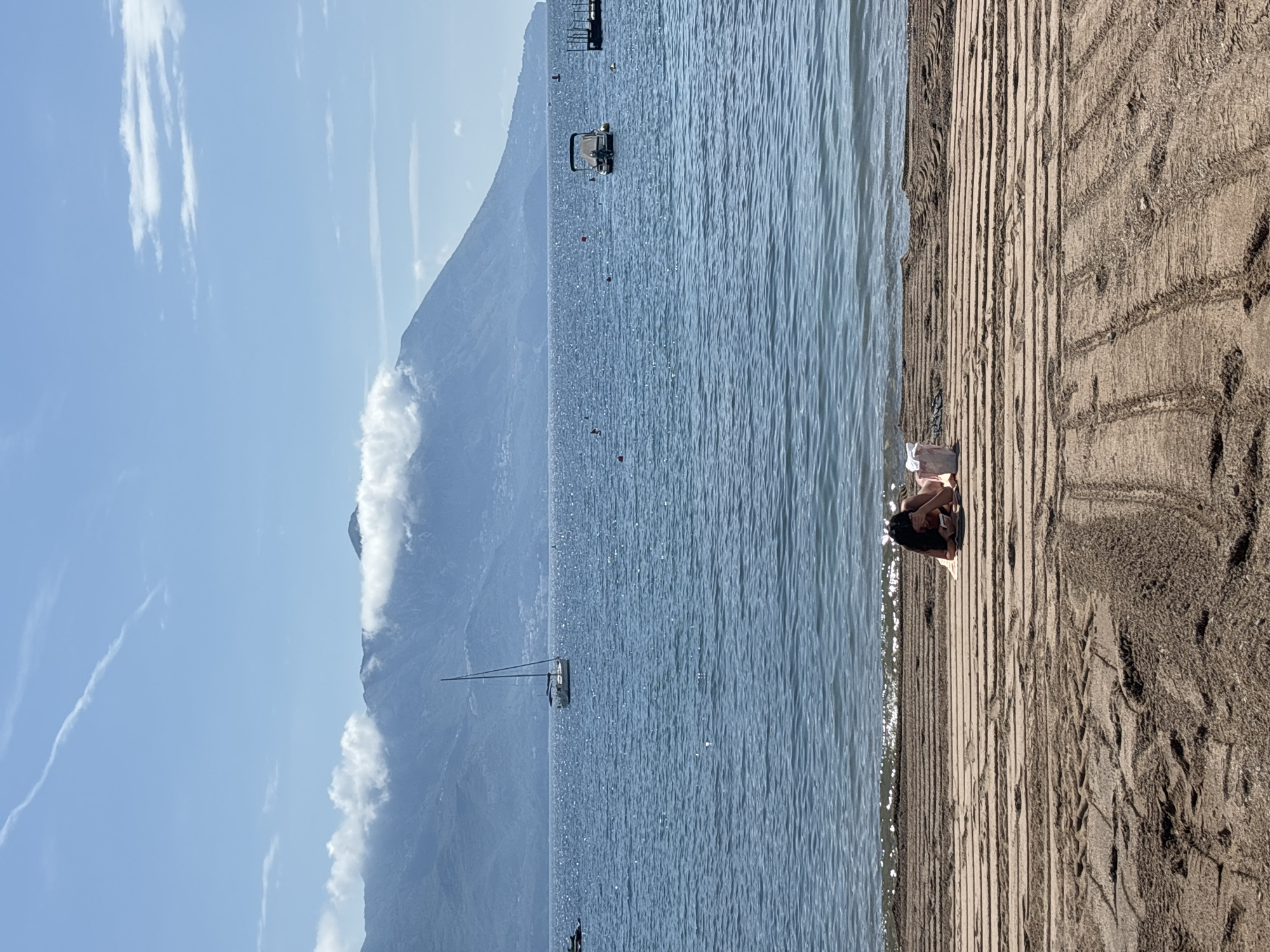 The image shows a person sitting on a wooden boardwalk beside a beach and the Adriatic Sea in Budva, Montenegro. The person has dark hair and is wearing a dark top or dress. They appear to be holding a book or device. The boardwalk leads to a sandy beach area with footprints. The sea is calm with a few boats in the distance. A large mountain range is visible in the background, partially obscured by clouds. The sky is blue with scattered clouds. The overall lighting suggests it is daytime.