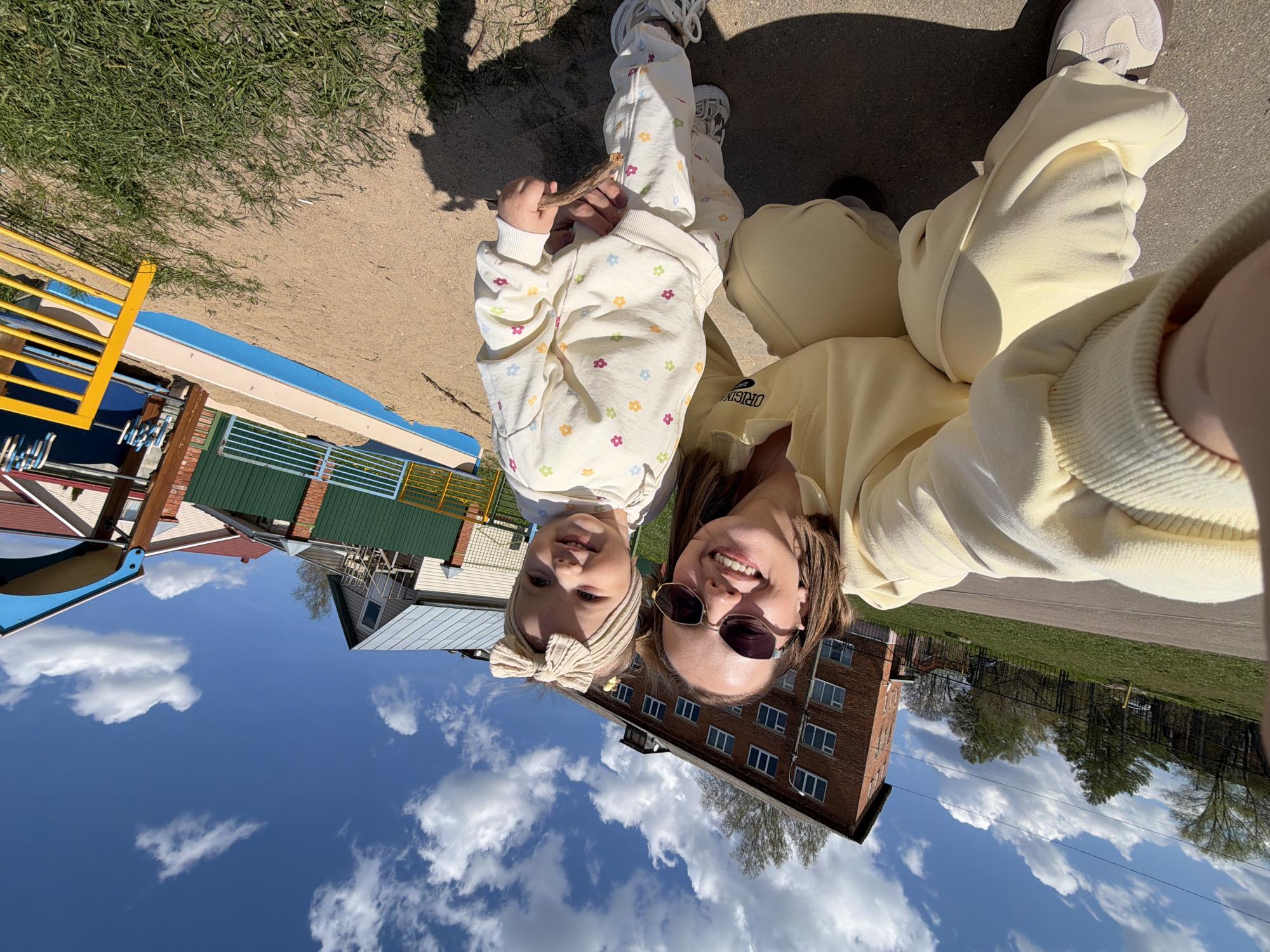 A selfie shows a woman with sunglasses and a young child outdoors on a sunny day in Reshetnikovo, Russia. The woman wears a yellow outfit. The child wears a patterned white outfit. A playground and brick building are in the background, along with a blue sky with white clouds. The ground is a mix of asphalt and sand.