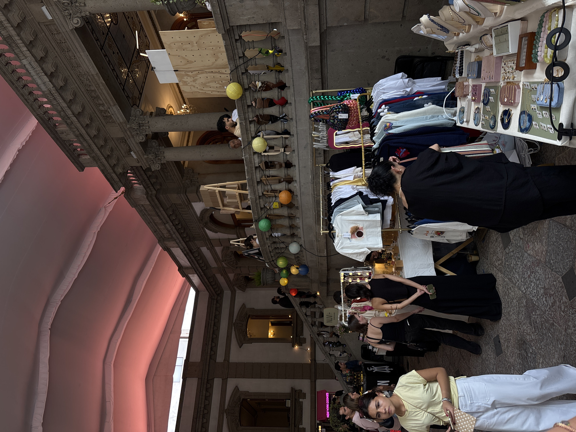 The image captures an interior courtyard scene in Mexico City, Mexico, which has been transformed into an open-air market. The space is enclosed by tall, ornate stone architecture featuring columns and arched openings. A pink awning covers part of the courtyard, casting a warm hue. Stalls display clothing and jewelry. Several individuals are browsing the merchandise, while others interact with vendors. Neon signs add a modern touch to the space.