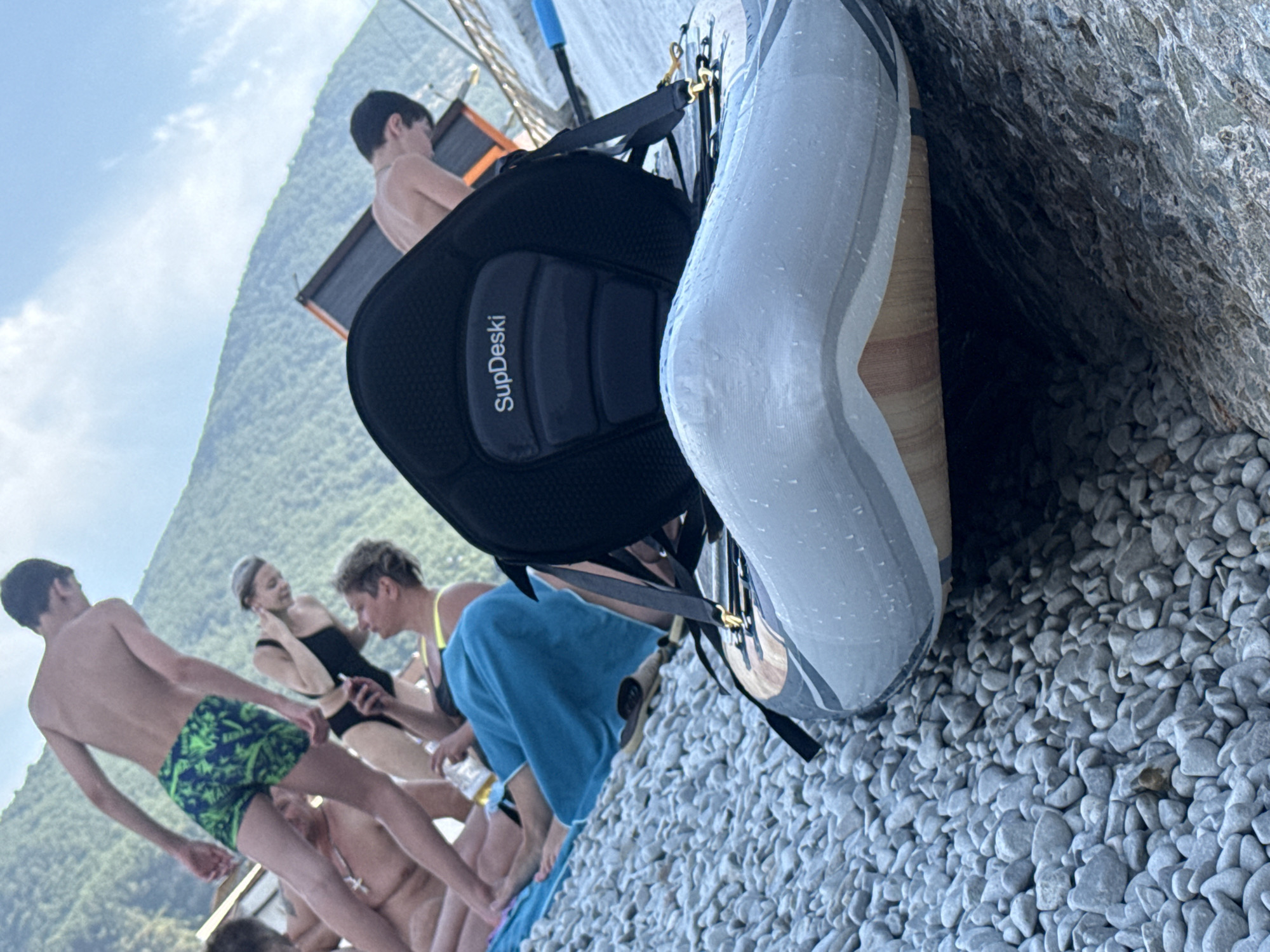 In Partenit, Ukraine, a group of people are gathered on a pebble beach. In the foreground, a paddleboard with a black seat labeled "SupDeski" rests on the stones. Behind it, a young man with dark hair wears green and blue patterned swim trunks. To his left, a woman in a black swimsuit touches her hair, and another person is seated with a blue towel. In the background, a mountainous landscape is visible under a partly cloudy sky.