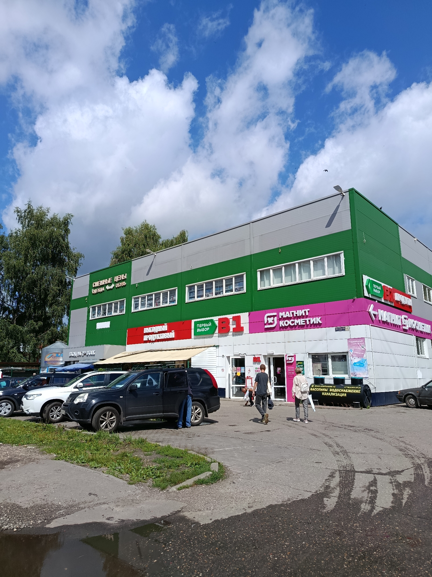 The image shows a retail building in Orekhovo-Zuyevo, Russia, on a cloudy day. The building has a green and gray facade with several storefronts. A large "Magnit Kosmetik" sign is visible, along with other signs indicating various shops, including "Odezhda Obuv" (Clothing Shoes) and "Pervyy Vybor" (First Choice). Several cars are parked in front of the building, and a few people are walking in the area. The ground in front of the building is paved, with some grass growing on the side and a puddle of water in the foreground. The sky is blue with scattered white clouds.