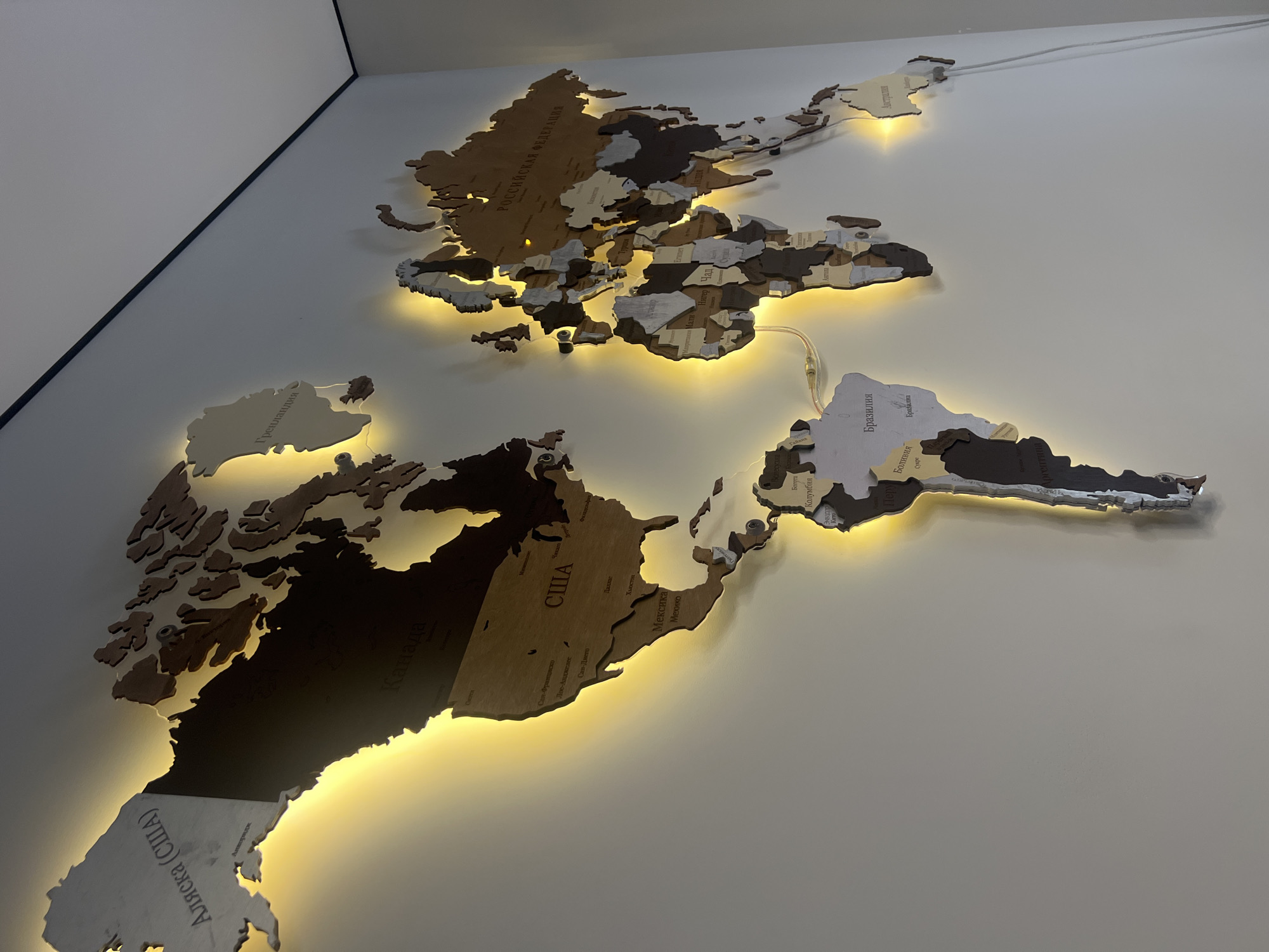 This is a well-lit, close-up, eye-level shot of a wooden world map art piece mounted on a white wall. The map features various continents and countries intricately cut and assembled, with some sections being lighter wood and others darker. The pieces are outlined with a warm yellow LED light that casts a glow around the edges. Several countries and cities are labeled in Cyrillic script. Prominently visible labels include "Аляска (США)" (Alaska, USA), "Канада" (Canada), "США" (USA), "Мексика" (Mexico), "Российская Федерация" (Russian Federation), "Австралия" (Australia), "Бразилия" (Brazil), and "Аргентина" (Argentina). Some of the cities labeled within the countries include "Сан-Франциско" (San Francisco), "Лос-Анджелес" (Los Angeles), "Сан-Диего" (San Diego), "Хьюстон" (Houston), "Даллас" (Dallas), "Москва" (Moscow), "Екатеринбург" (Yekaterinburg), "Кишинев" (Chisinau), "Анкара" (Ankara), "Каир" (Cairo), "Нагер" (N'Djamena), "Мали" (Mali), "Нджамена" (N'Djamena), "Судан" (Sudan), "Аддис-Абеба" (Addis Ababa), "Богота" (Bogota), "Колумбия" (Colombia), and "Лима" (Lima). There are visible wires connected to the LED lights, one of which snakes down the wall to the right. The wall appears clean and the room is dimly lit except for the illumination from the map's LEDs. The top right corner of the image shows a thin black trim line where the wall meets the ceiling. There are no people or animals present.