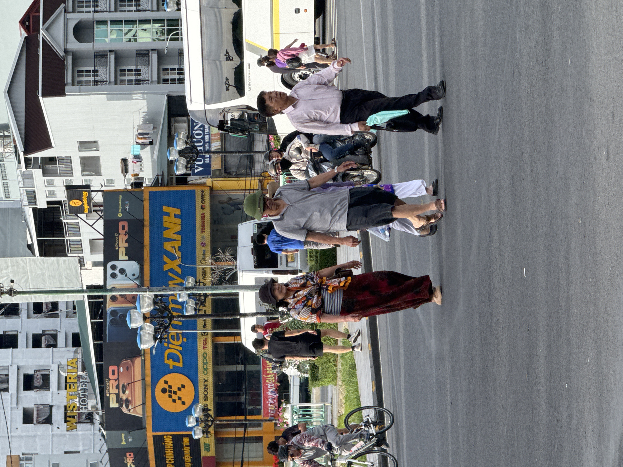 This is a street-level, medium-shot photograph taken in Da Lat, Vietnam, depicting a busy intersection with several individuals walking and riding. In the foreground, a woman wearing a wide-brimmed hat and a vibrantly patterned scarf over a striped shirt and a long red skirt walks across the street. To her left, a man in a gray t-shirt and black shorts walks with his head turned towards the right, carrying a light blue bag. Behind them, a man in a light pink collared shirt and black pants walks to the right, also carrying a bag. Further in the background, a person on a motorcycle, wearing a helmet and a light brown jacket, is visible. On the right side of the frame, a white bus with a yellow stripe is parked, and two individuals, one in a purple top and white shorts and another in a pink jacket and white shorts, are walking away from it. A man in a blue t-shirt walks further into the background. A cyclist in a grey and pink jacket is on the left side of the image, riding a white bicycle. The background shows buildings, including a white building with the "WISTERIA HOTEL" sign and a large blue and yellow sign for "Điện máy XANH", which is a Vietnamese electronics retailer. Streetlights with multiple bulbs are present along the roadside. The scene appears to be during daylight hours.