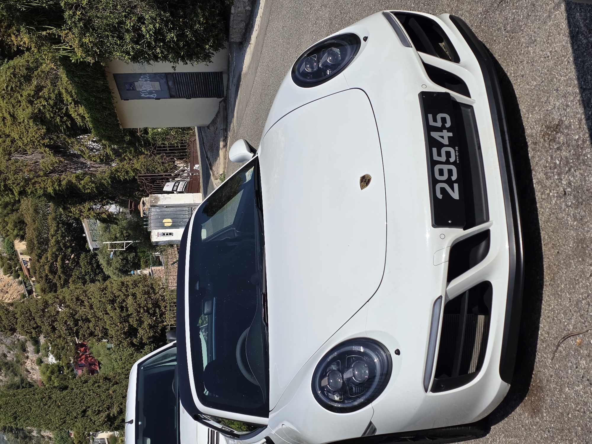 A white convertible Porsche 911 is parked on a paved road in Roquebrune-Cap-Martin, France. The car is facing forward, with its black convertible top down. The license plate reads "29545". To the left of the Porsche, a white van is partially visible. The background consists of a steep hillside covered in trees and houses. A small, light-colored building with a dark metal door and graffiti is located to the right of the car. A wrought-iron gate is visible behind the van. The scene is illuminated by sunlight, casting shadows on the road.