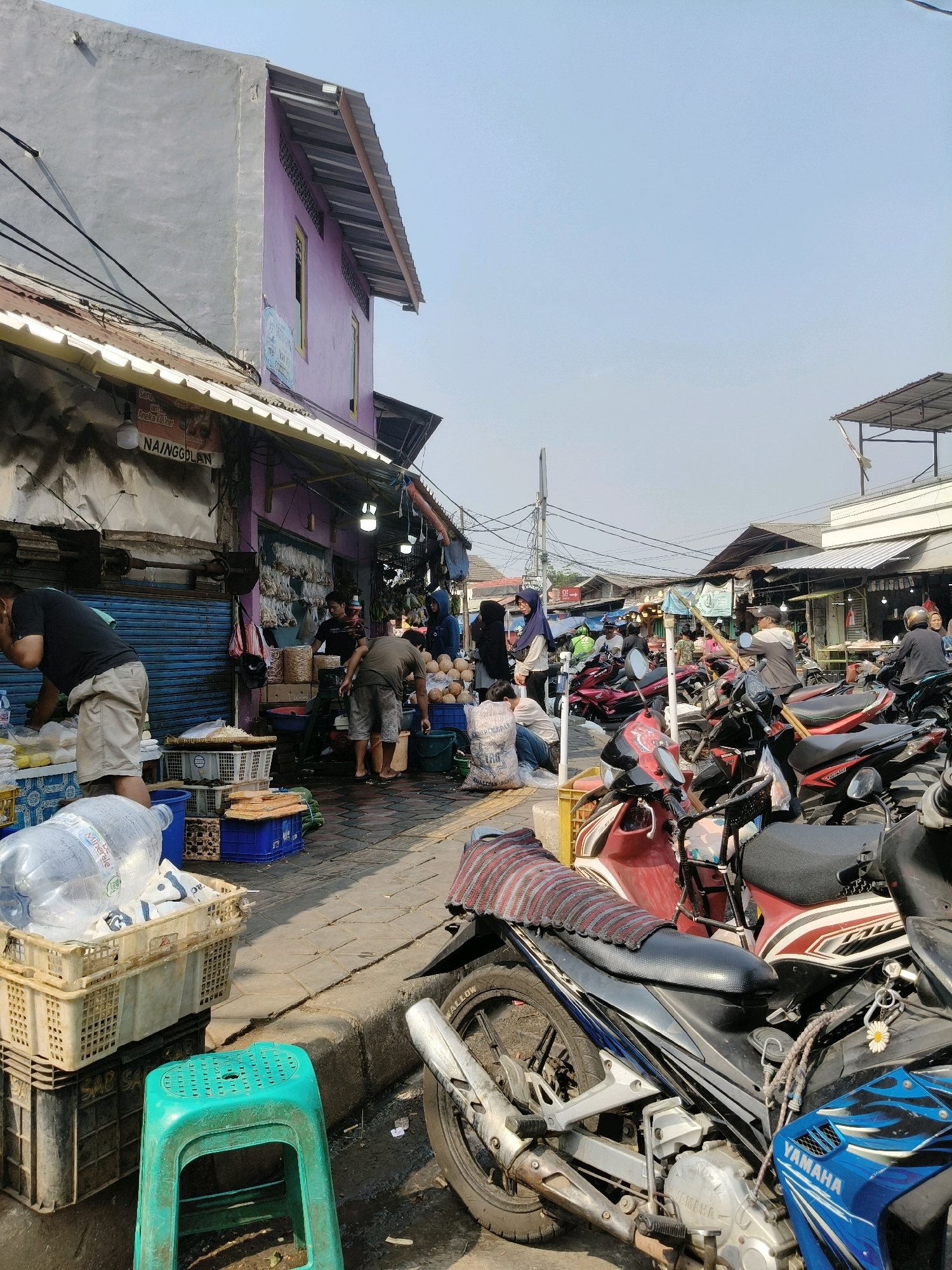 The image depicts a busy street market scene in South Tangerang, Indonesia. In the foreground, a turquoise plastic stool is placed on the ground next to several stacked crates, one of which holds a large bottle of mineral water. Partially visible is the front of a blue and black Yamaha motorcycle. The street is paved with cobblestones and shows signs of being wet.
Further into the scene, a row of motorcycles, primarily red and black, are parked on the side of the street, some with riders. The market stalls lining the street are covered by awnings and corrugated metal roofs. One stall, painted purple, has a sign reading "NAINGGOLAN" above the entrance. Various goods, including what appear to be coconuts, dried goods in bags, and produce, are displayed for sale. Several people are engaged in market activities, such as buying, selling, and interacting. The overall atmosphere suggests a typical bustling Indonesian market day. The sky is clear and bright, indicating daytime.