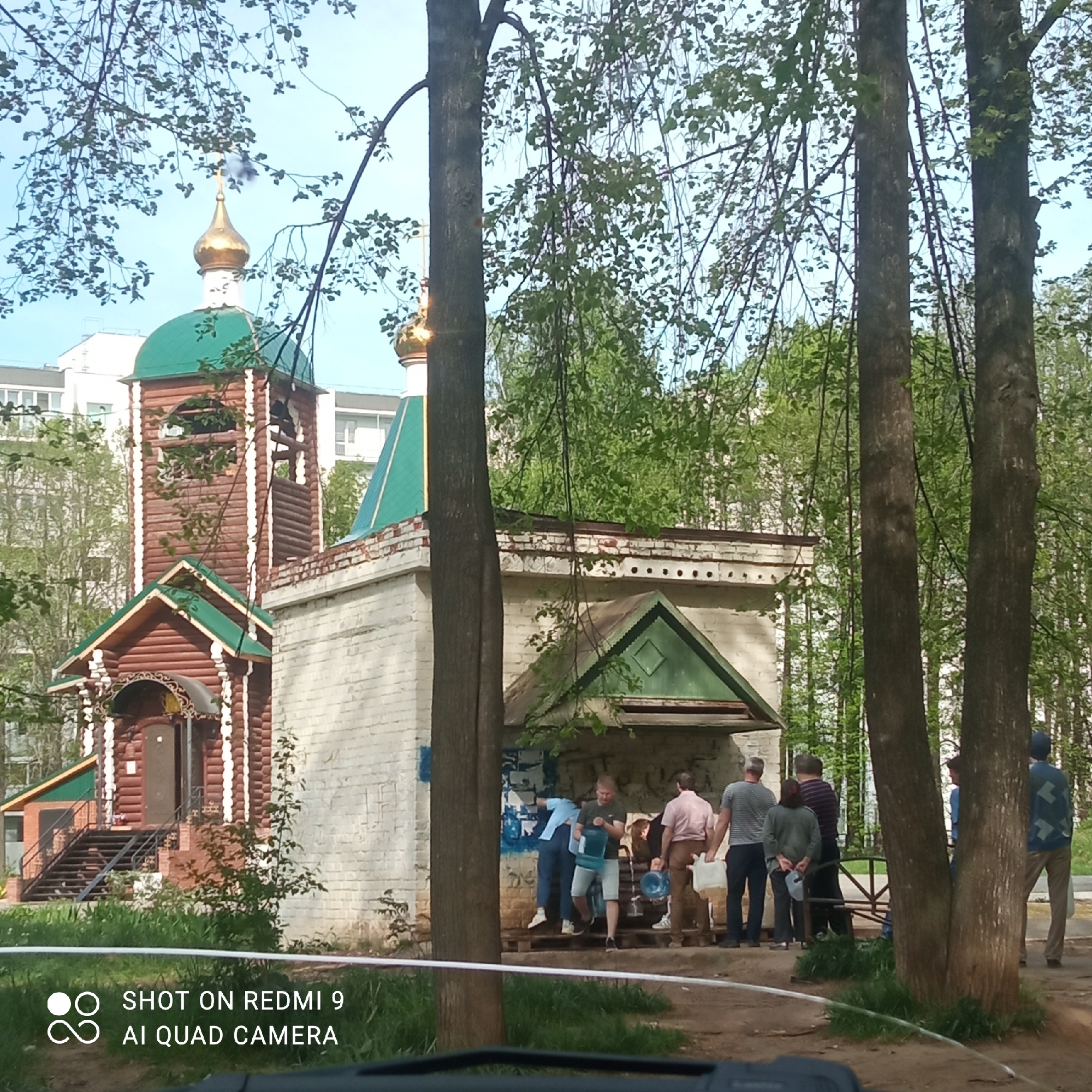 The image shows a crowd of people gathering at the Sacred Spring of St. George the Victorious, located in Chaikovsky, Perm Krai, Russia. The spring is housed in a small, white-brick structure adjacent to a wooden church. Several people are filling containers with water from the spring. The church has a wooden bell tower and a green roof with golden domes. The scene is partially obscured by trees in the foreground. A residential building can be seen in the background.