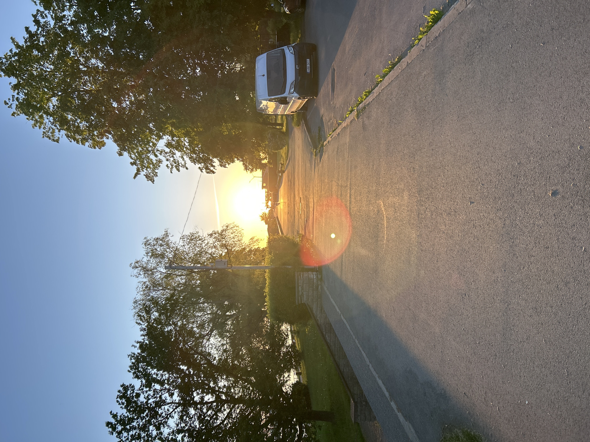 The image depicts a wide asphalt promenade or path during a sunset or sunrise in Tallinn, Estonia. The low-lying sun, a bright orange-yellow orb, is positioned centrally above the horizon, casting a strong backlight that illuminates the scene with warm, golden light and creates long shadows extending towards the viewer. A prominent red-orange circular lens flare is visible in the lower-middle portion of the image.
On the left side of the path, a grassy bank slopes downwards towards a body of water, likely a bay or the Baltic Sea. Dark green trees with dense foliage line this bank, appearing silhouetted against the bright sky. A low stone wall structure separates the path from the grassy area. In the middle distance on the path, several human figures are visible as dark silhouettes, walking towards the setting sun.
The right side of the path is bordered by more trees and dense green bushes. A white panel van, identifiable as a Fiat Ducato or similar model, is parked on the right shoulder, facing the camera. Its Estonian license plate, "488 SCT", is clearly visible. A portion of another dark vehicle is visible behind the van. A utility pole with attached equipment stands behind the van and trees.
The asphalt path itself is grey, with its texture visible. A single white line is painted along its left edge. Small patches of green vegetation are growing along the right edge of the path where it meets the shoulder. The sky above is clear and light blue, transitioning to orange and yellow closer to the horizon where the sun is located.