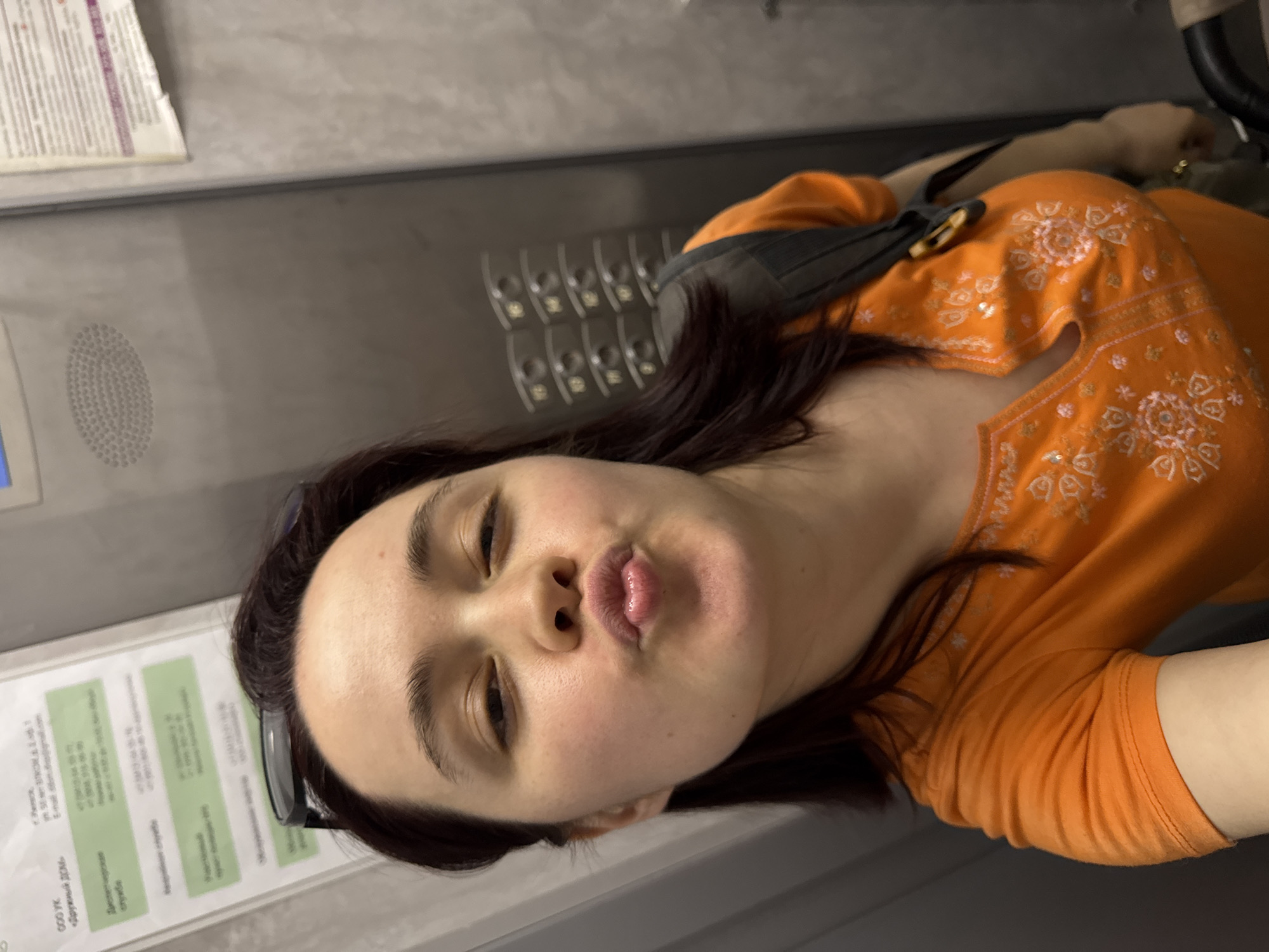 A woman is posing for a selfie in what appears to be the lobby of a building in Izhevsk, Russia. She is wearing an orange top with a floral pattern and a black strap of a bag is visible over her shoulder. Behind her, there is a wall with a series of key slots or lockers, as well as a bulletin board with printed information in Russian. The background appears to be the interior of a public or commercial space.