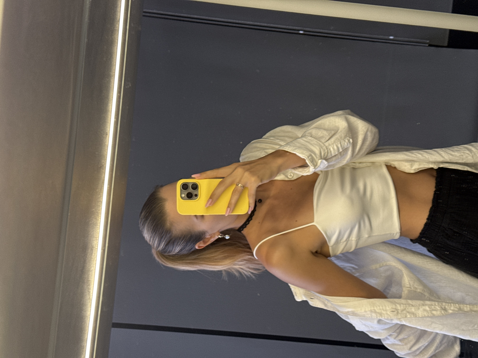 A woman is reflected in a mirror, holding a yellow smartphone in her right hand. She is wearing a white camisole, a white collared shirt, and black pants. She has blonde hair. The background is dark gray. The location appears to be a dressing room with an illuminated mirror.