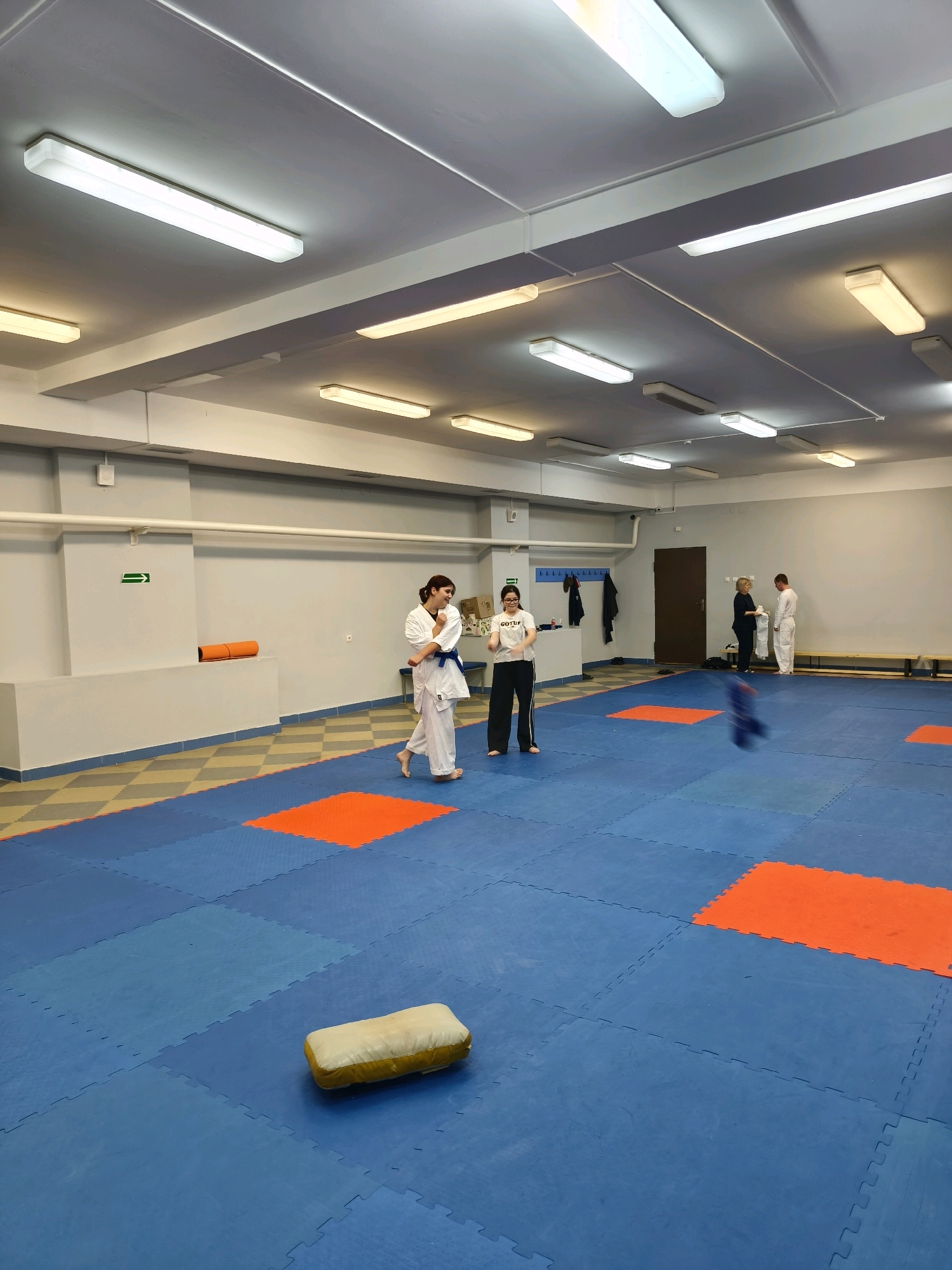 The image depicts an indoor martial arts training hall. The floor is covered with blue interlocking mats, interspersed with orange square mats. In the foreground, a light-colored, cylindrical training pillow lies on the blue mats. In the mid-ground, two young women in white martial arts uniforms are practicing. One is in motion, facing away from the camera, while the other stands facing the camera, holding her hands in a defensive stance. Further in the background, on the right side of the image, an adult woman in dark clothing is handing a white garment to a man in a white martial arts uniform. The hall has a high ceiling with multiple fluorescent light fixtures. The walls are painted in muted tones of gray and blue, with some shelving and a doorway visible. The environment appears clean and organized, suitable for martial arts training. The location is identified as Karachayevsk, Russia.