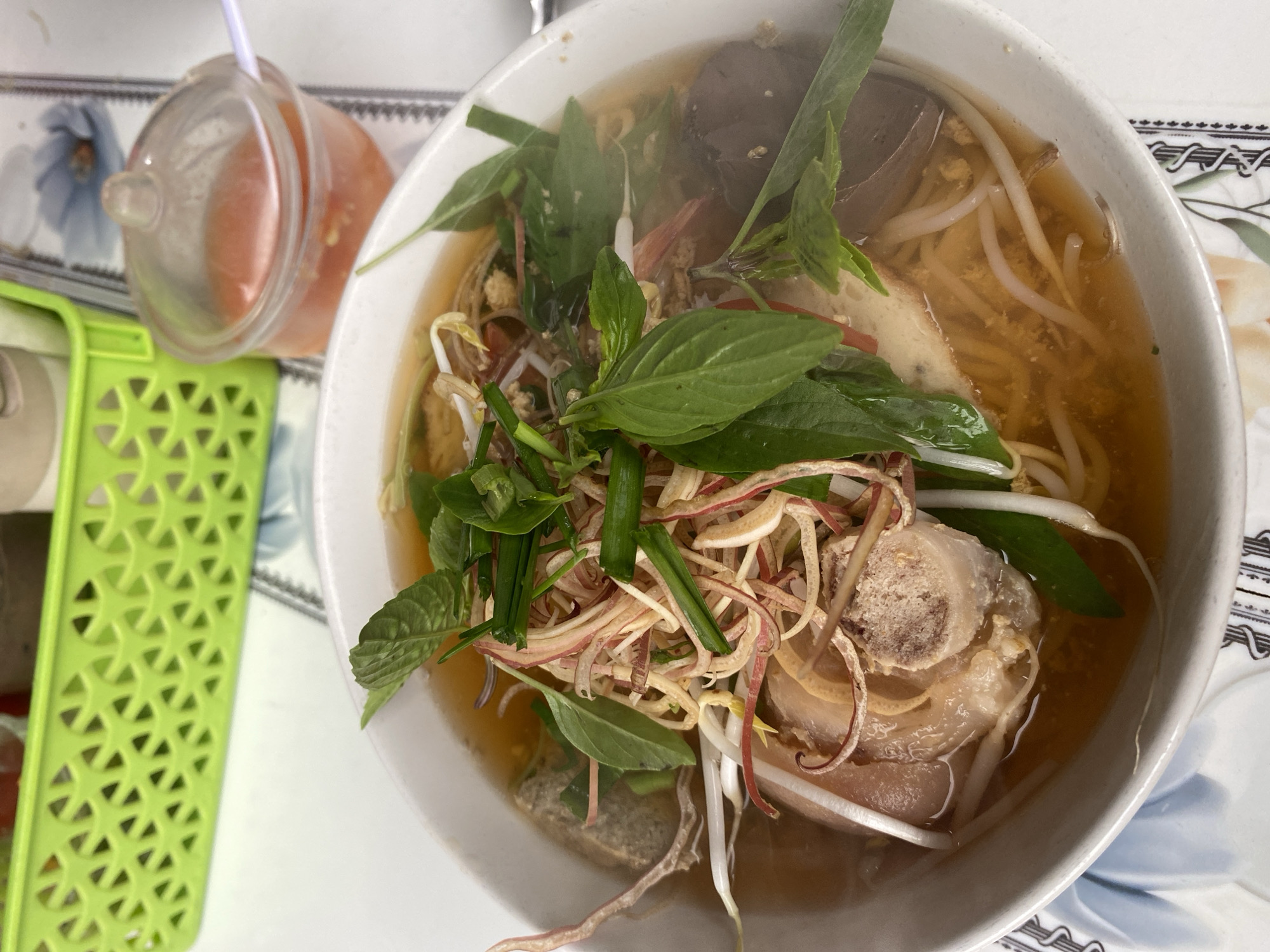 An overhead medium shot captures a close-up of a bowl of Vietnamese pho soup, served on a table with a patterned tablecloth. The soup features thin noodles submerged in a clear broth, topped with a generous garnish of fresh green herbs, bean sprouts, thinly sliced pink pickled ginger, and a large piece of what appears to be braised pork knuckle with bone. Some slices of processed meat and what looks like a piece of blood cake are also visible. To the upper right of the bowl, a small plastic container holds a reddish dipping sauce, with a white spoon emerging from it. Behind the soup, a bright green plastic basket with an intricate lattice pattern is visible, containing various condiments. The environment suggests a casual dining setting in an unknown urban location.