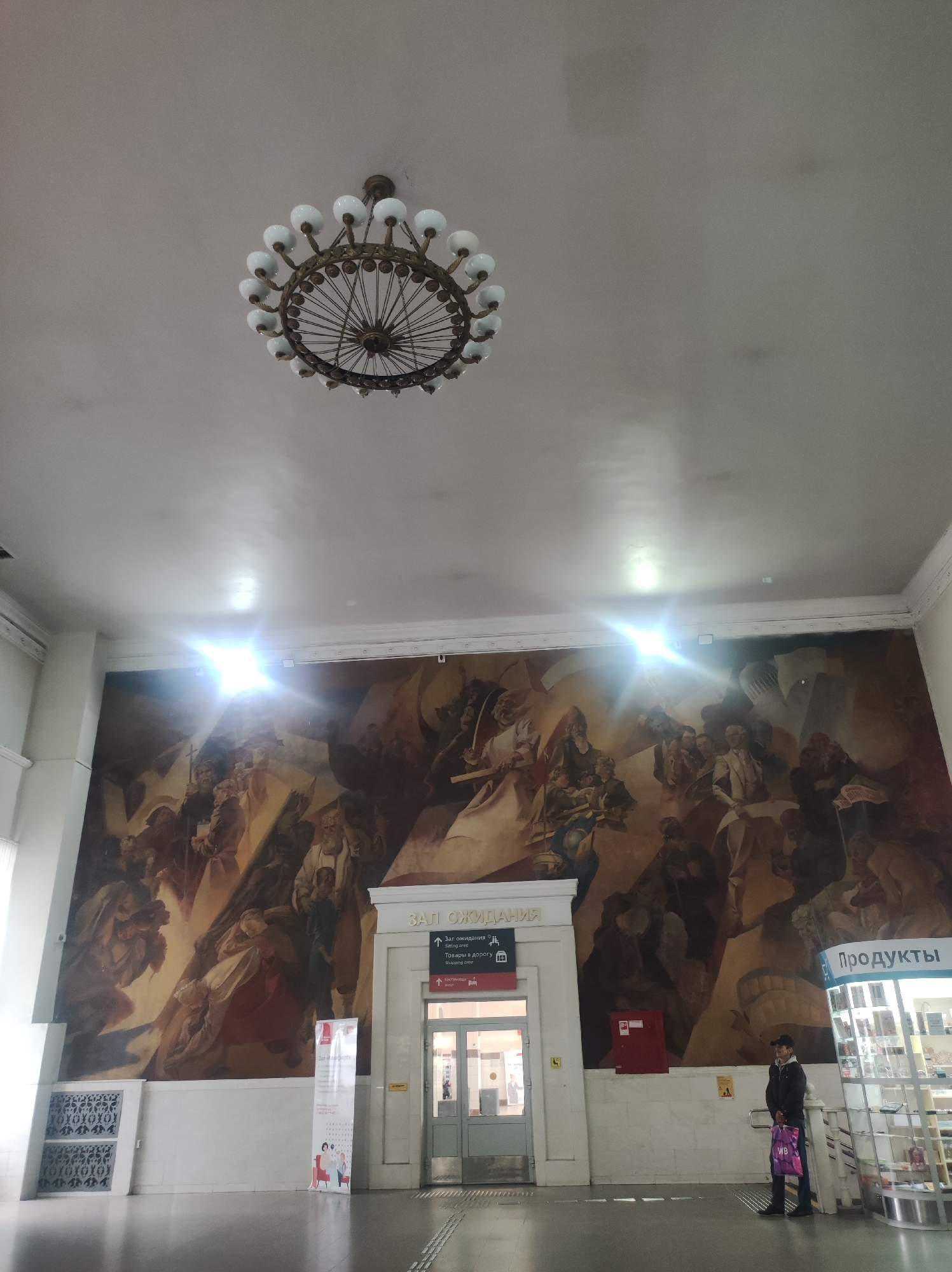 The image depicts the interior of a building, possibly a train station or public hall, with a large mural covering the wall. In the foreground, a man stands near a retail display labeled "ПРОДУКТЫ" (Products). To his left is an entrance marked "ЗАЛ ОЖИДАНИЯ" (Waiting Room). Above, a large, multi-bulb chandelier hangs from the ceiling. The mural itself is a dramatic, figurative scene with multiple figures in dynamic poses. The overall lighting suggests an indoor, possibly well-lit environment. This scene is located in the city of Kirov, Russia, at the Kirov railway station.