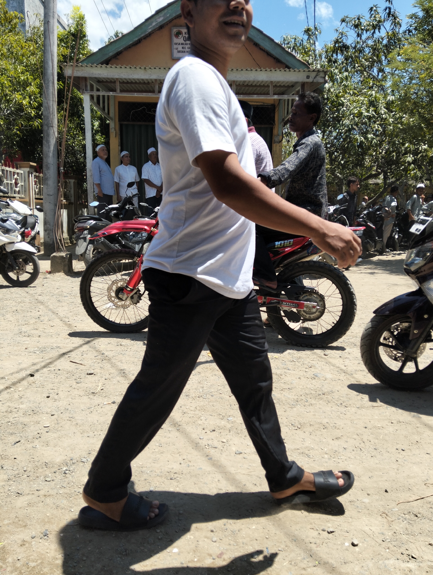 In Teungoh, Indonesia, a man in a white t-shirt and black pants walks on a dirt road in front of a building. The building appears to be a small mosque or community center, with a sign on it that reads "KUA Q". Several other people, some wearing white skullcaps, stand near the building. Motorcycles are parked on the side of the road. The sky is blue with some clouds, and trees are visible in the background. The man in the foreground is wearing black sandals.