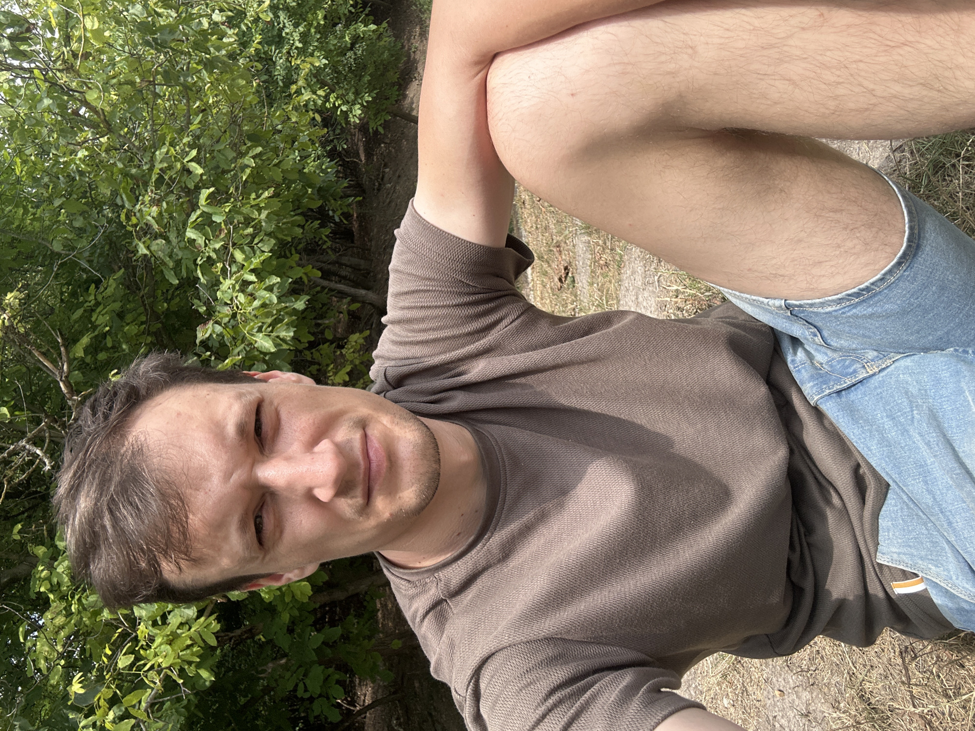 A Caucasian male, appearing to be in his late 20s or early 30s, is seated outdoors with trees and foliage in the background. He has short brown hair, light skin, and is wearing a brown t-shirt and blue denim shorts. His left arm is bent and resting on his left knee, which is crossed over his right leg. He is looking towards the camera with a neutral expression. The ground beneath him appears to be dry earth and some sparse grass. The image is a close-up shot, focusing on the man's upper body and legs, with the natural environment forming a blurred backdrop. The location is identified as Makiv, Ukraine.