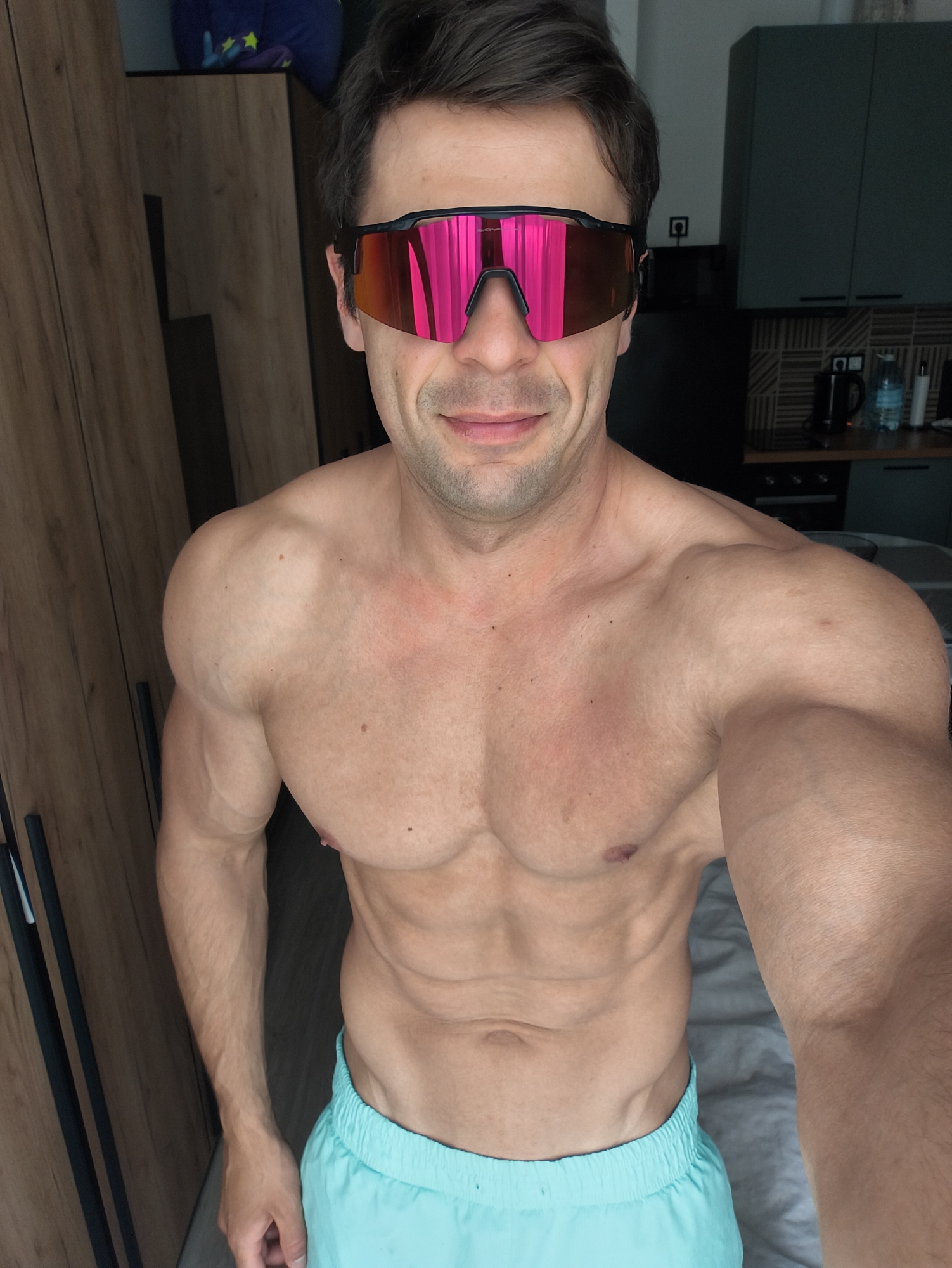 A shirtless man wearing sunglasses and light blue shorts is standing indoors. There are closed wooden cabinets and a kitchen visible in the background. A kettle and a water bottle are on the kitchen counter.