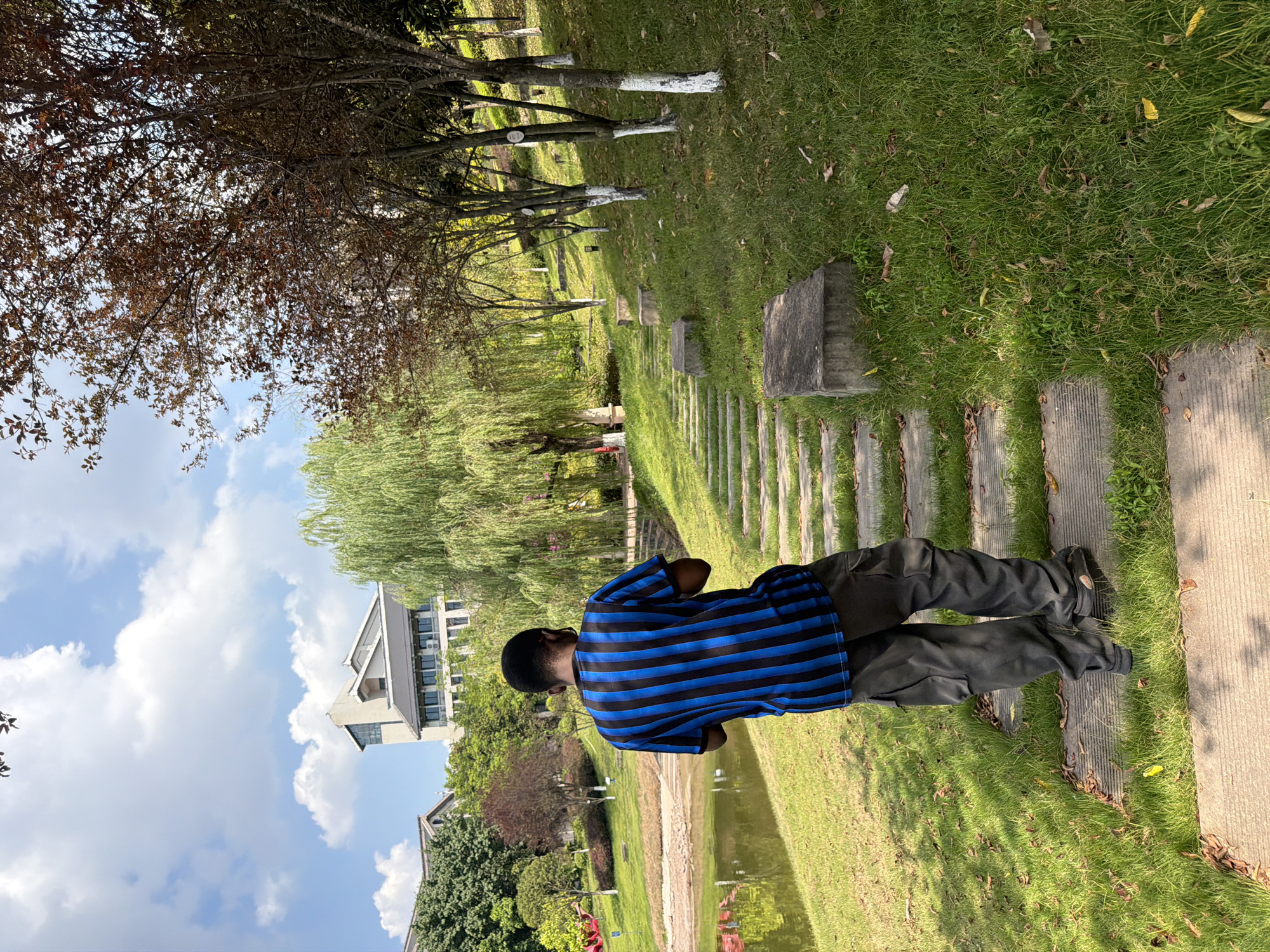 A lone Black male, wearing a blue and black striped t-shirt and dark cargo pants, walks away from the camera on a stone pathway through a grassy park. The pathway curves to the right as it ascends a gentle slope lined with trees. The trees on the right have white-painted trunks and some have reddish-brown leaves, while a weeping willow tree with vibrant green foliage is visible on the left. In the background, a body of water, likely a pond or lake, reflects the sky and surrounding greenery. Beyond the pond, a modern building with multiple stories and a distinctive angular roofline is visible, nestled amongst trees. The sky is blue with scattered white clouds, indicating a bright, sunny day. The scene appears to be a public park or university campus, possibly located in Jiangjin, China, given the architectural style and landscaping.