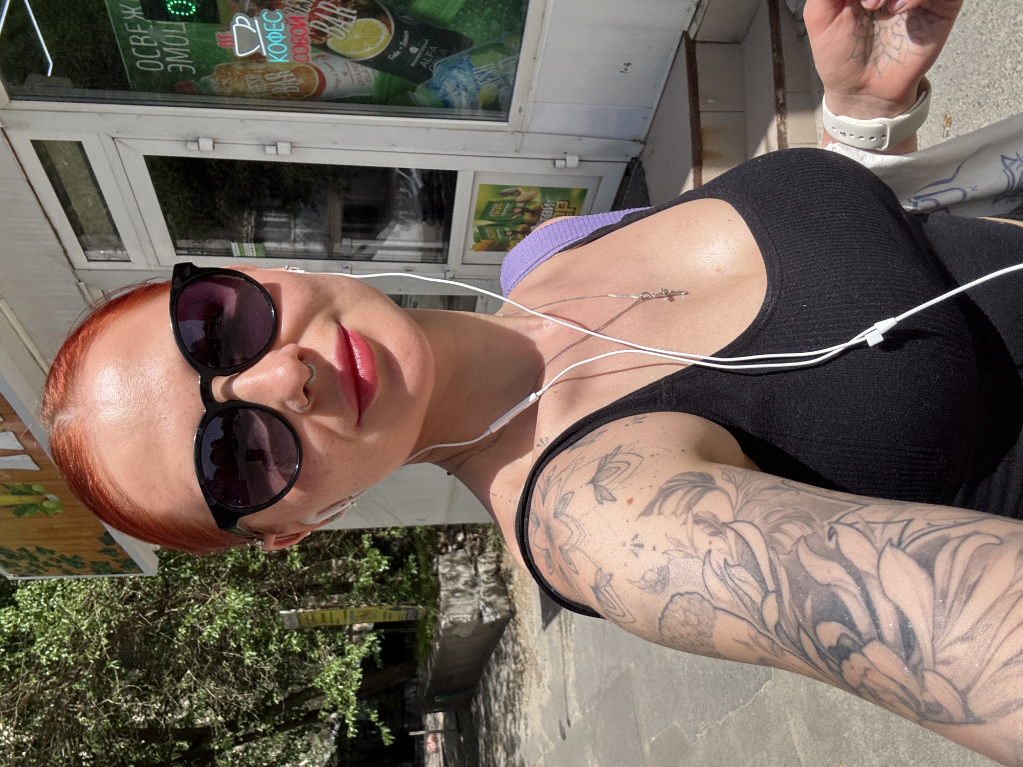 A selfie taken outdoors in Rostov-on-Don, Russia shows a fair-skinned woman with bright red hair and dark sunglasses, a nose ring, and a silver necklace with a cross. She is wearing a black ribbed top with a purple bra strap visible. Her left arm, extensively tattooed with a floral design, is visible. White earbuds extend from her ears. She is standing on a paved area. Behind her is the exterior of a shop with advertisements for coffee and other products visible through the glass door. Green foliage is visible to the left. A building facade and a sidewalk are also visible. Another hand is visible in the upper right corner of the image, with a white watch and a hand tattoo.