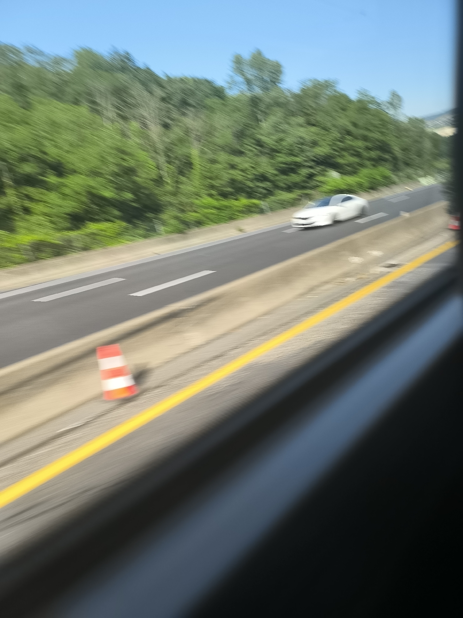 The image shows a view from inside a moving vehicle on the A47 autoroute, a motorway in France, specifically within the vicinity of L'Horme. The direction of travel seems to be southward, based on the placement of the traffic cone on the shoulder of the road. A white car is visible on the rightmost lane of the highway. Adjacent to the road is a line of green trees. The sky is clear and blue. A yellow lane marking divides the road from the right-hand shoulder. There is a red and white traffic cone located on the side of the road near the yellow line. White dashed lines mark the lanes on the highway.