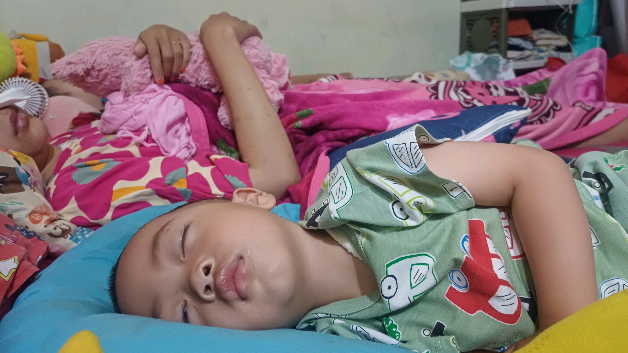 The image shows two young children and an adult lying on a bed. The foreground features a young child with closed eyes, sleeping on a blue pillow. The child is wearing a light green shirt with a pattern of cartoon vehicles. Behind this child, another child is visible, also appearing to be asleep, with their face partially obscured by blankets. This child is wearing a brightly colored shirt with a floral pattern. An adult's arm and hand are visible in the upper left corner, resting on a pink stuffed animal. The scene appears to be indoors, likely a bedroom, with blankets and pillows spread across the bed. The lighting suggests it could be daytime. The overall impression is one of a peaceful and sleepy environment. There is no visible text in the image.