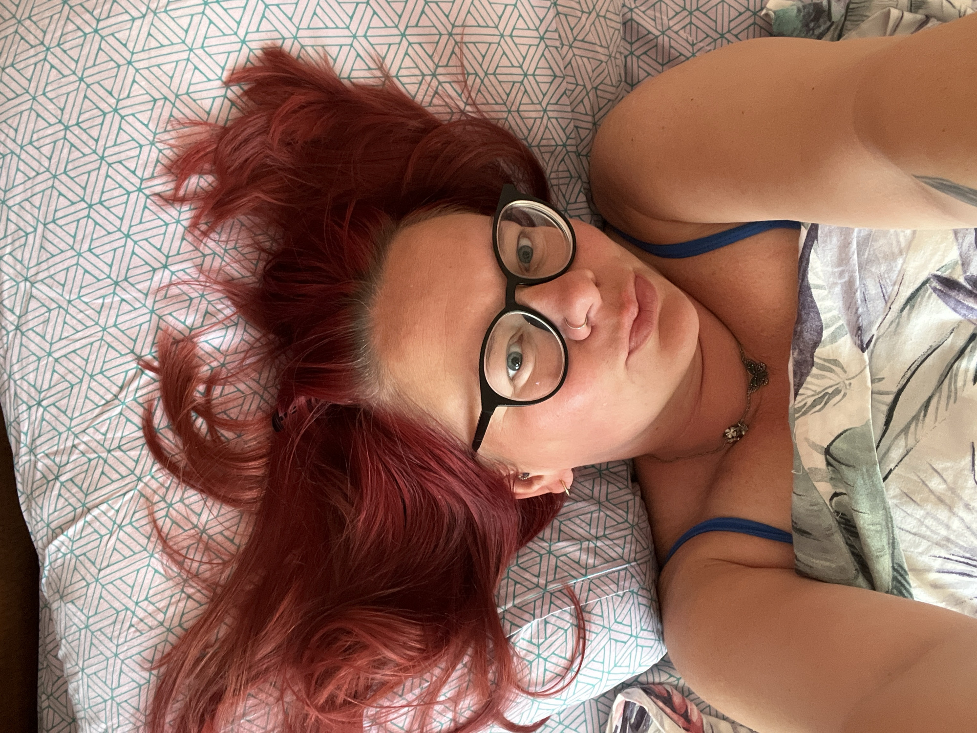 A woman with red hair and glasses lies on a patterned bedsheet. She is wearing a blue tank top. The location is unknown, and there is no information to suggest it is in Chaikovsky, Russia.