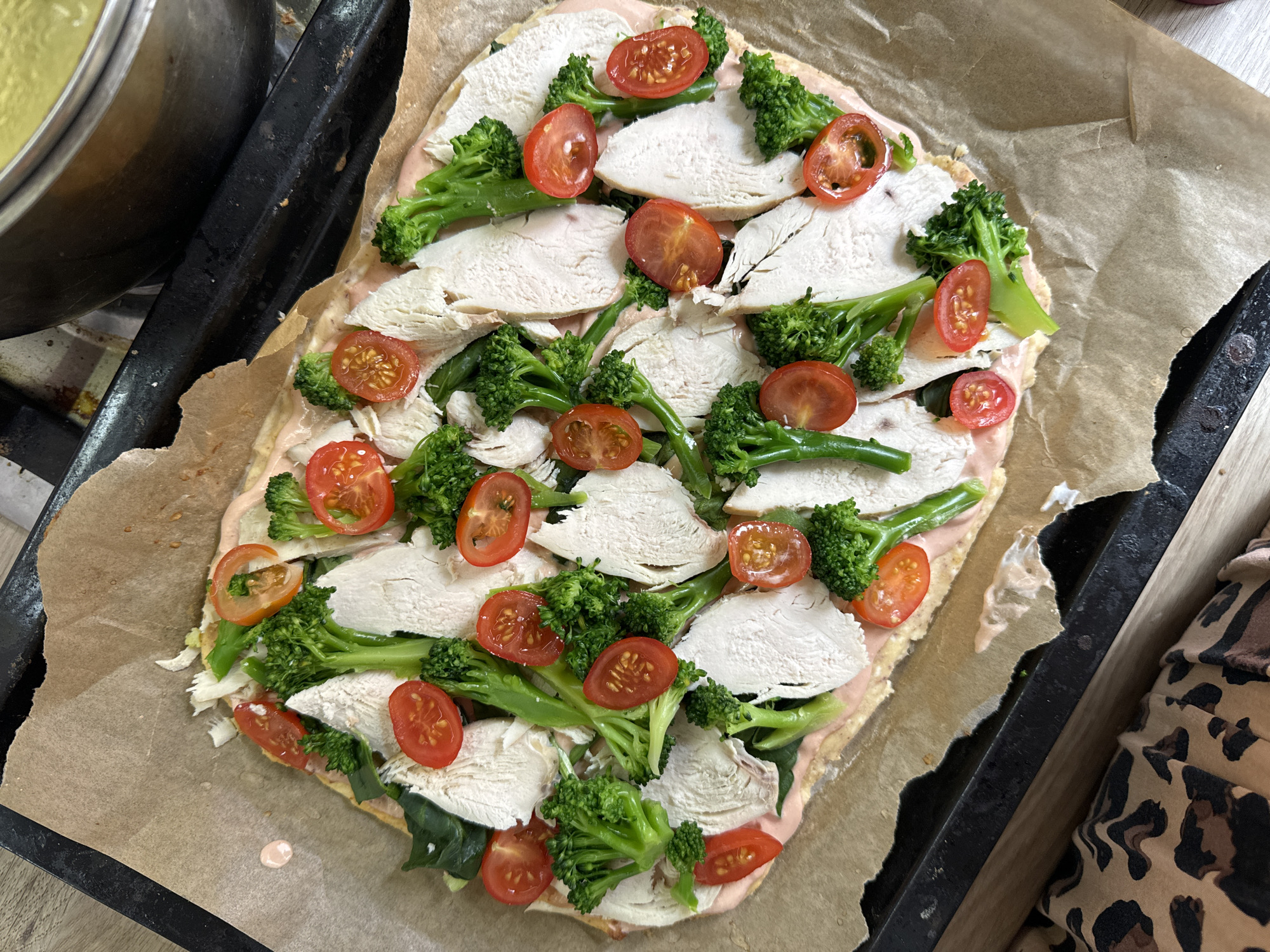A rectangular flatbread pizza, placed on baking parchment on a dark baking sheet, features a light pink sauce base, covered with layers of sliced white meat, broccoli florets, and halved cherry tomatoes. A silver saucepan is visible in the top left corner, while a portion of leopard-print clothing is present in the bottom right corner. The scene appears to be indoors, likely in a kitchen. The location is not possible to determine from the image.